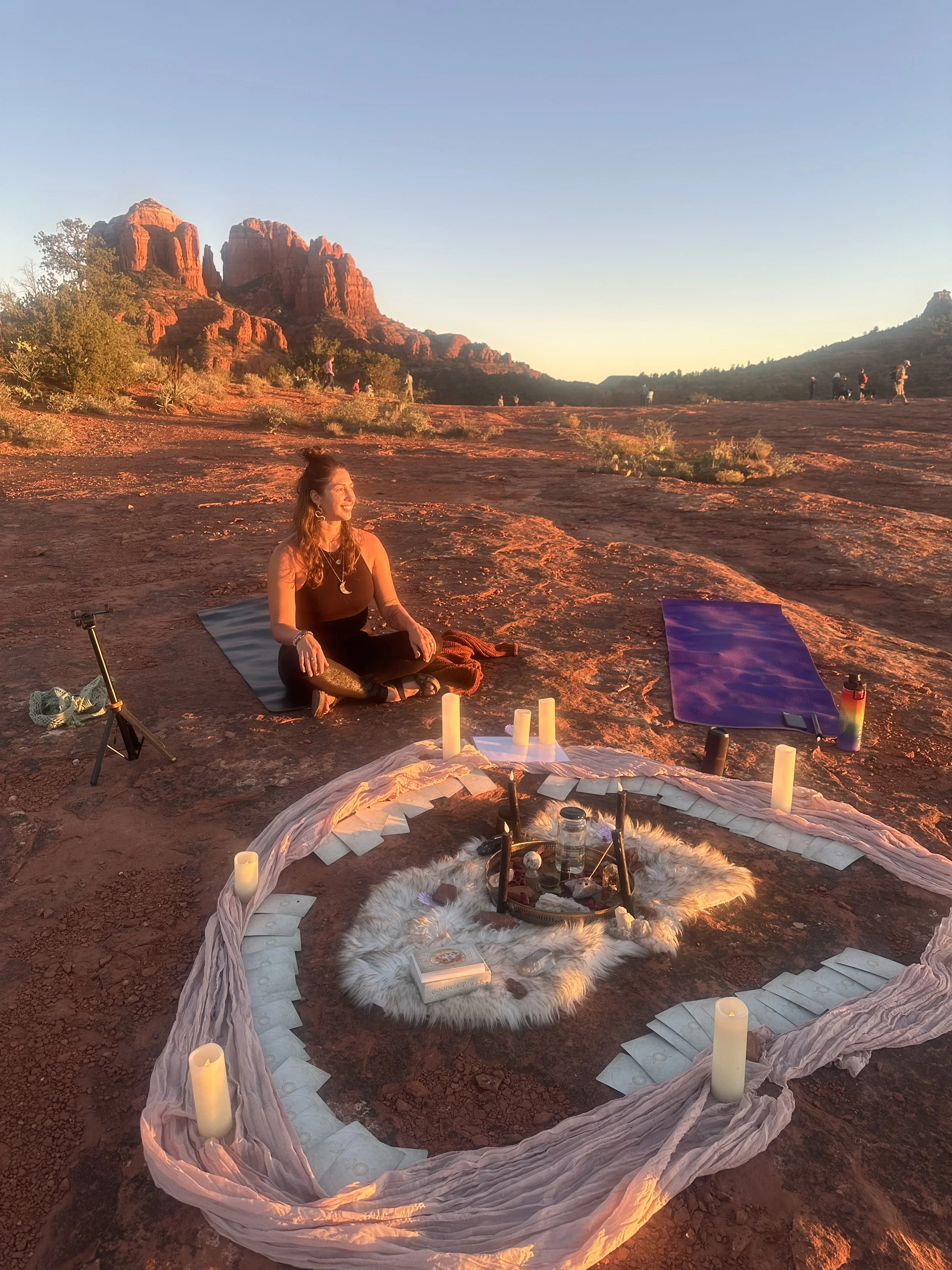 A woman practicing yoga outdoors in a desert landscape with red rock formations, during sunset. She is sitting on a yoga mat near a circular arrangement of candles and objects on a fluffy white rug, with additional yoga mats nearby.