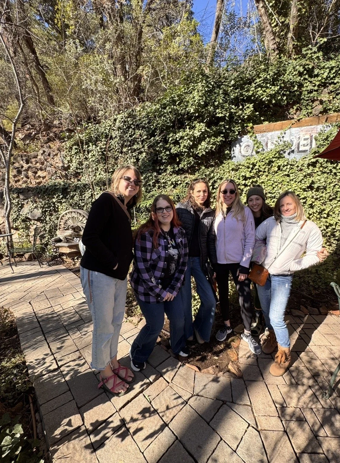 Group of six women smiling outdoors on a paved patio with greenery and a stone wall in the background.