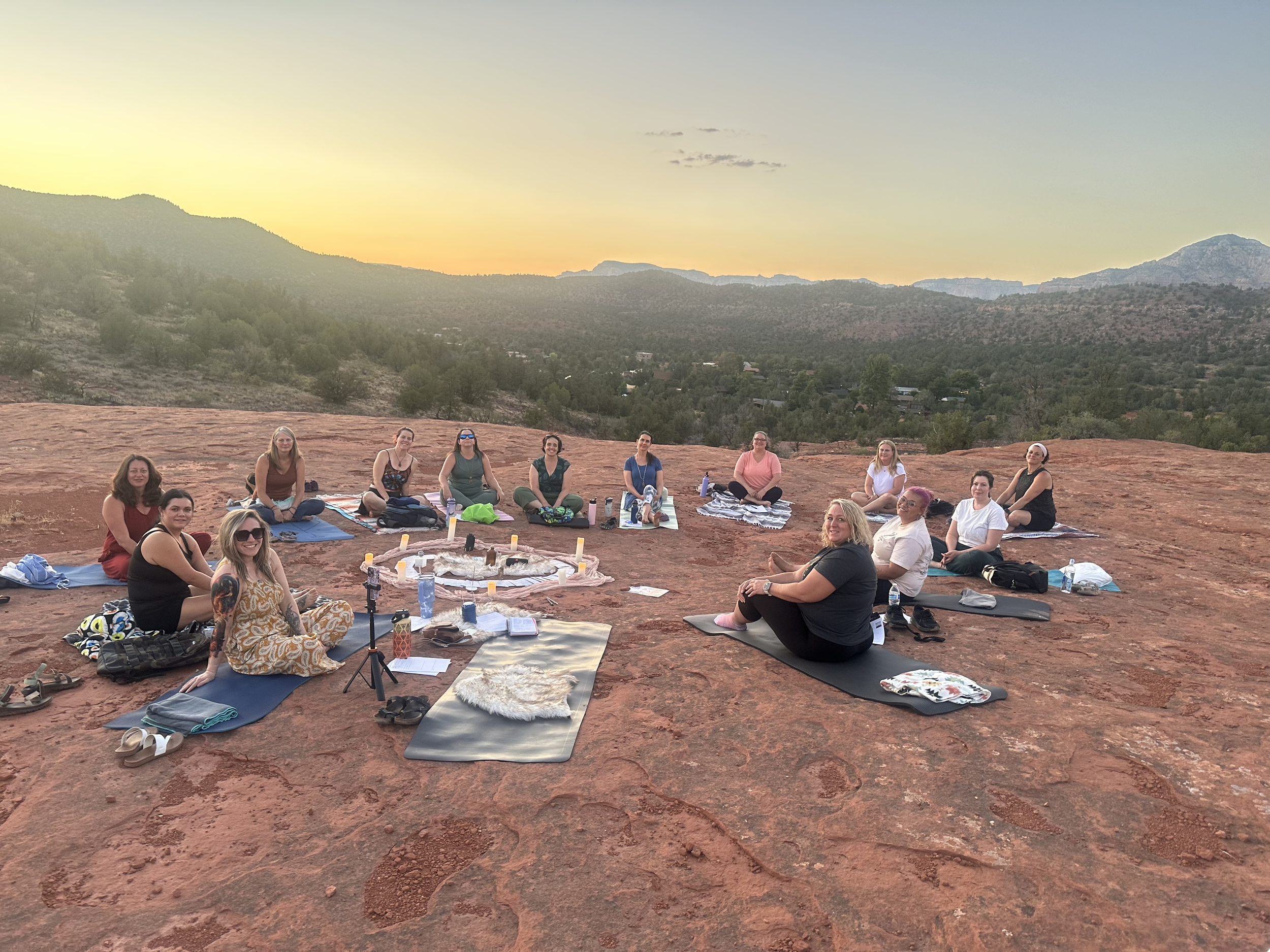 Group of women practicing yoga outdoors on a rocky terrain during sunset, sitting on yoga mats in a semi-circle with scenic mountains in the background.