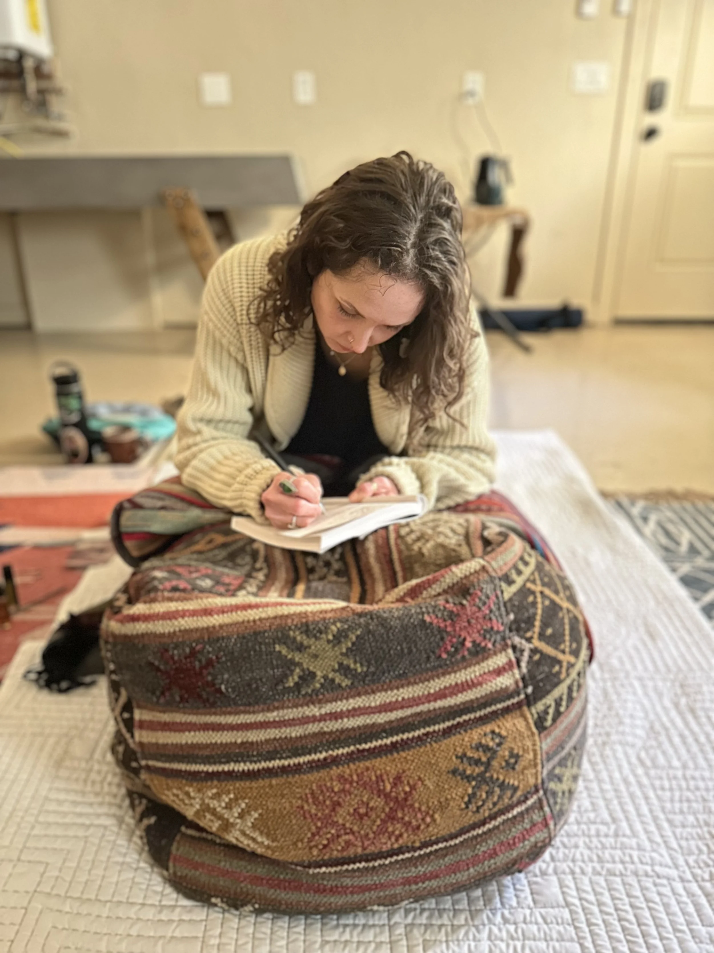 A woman with curly hair writing or drawing on a notepad, sitting cross-legged on rounded pillows with intricate patterns, on a quilted blanket in a cozy room.