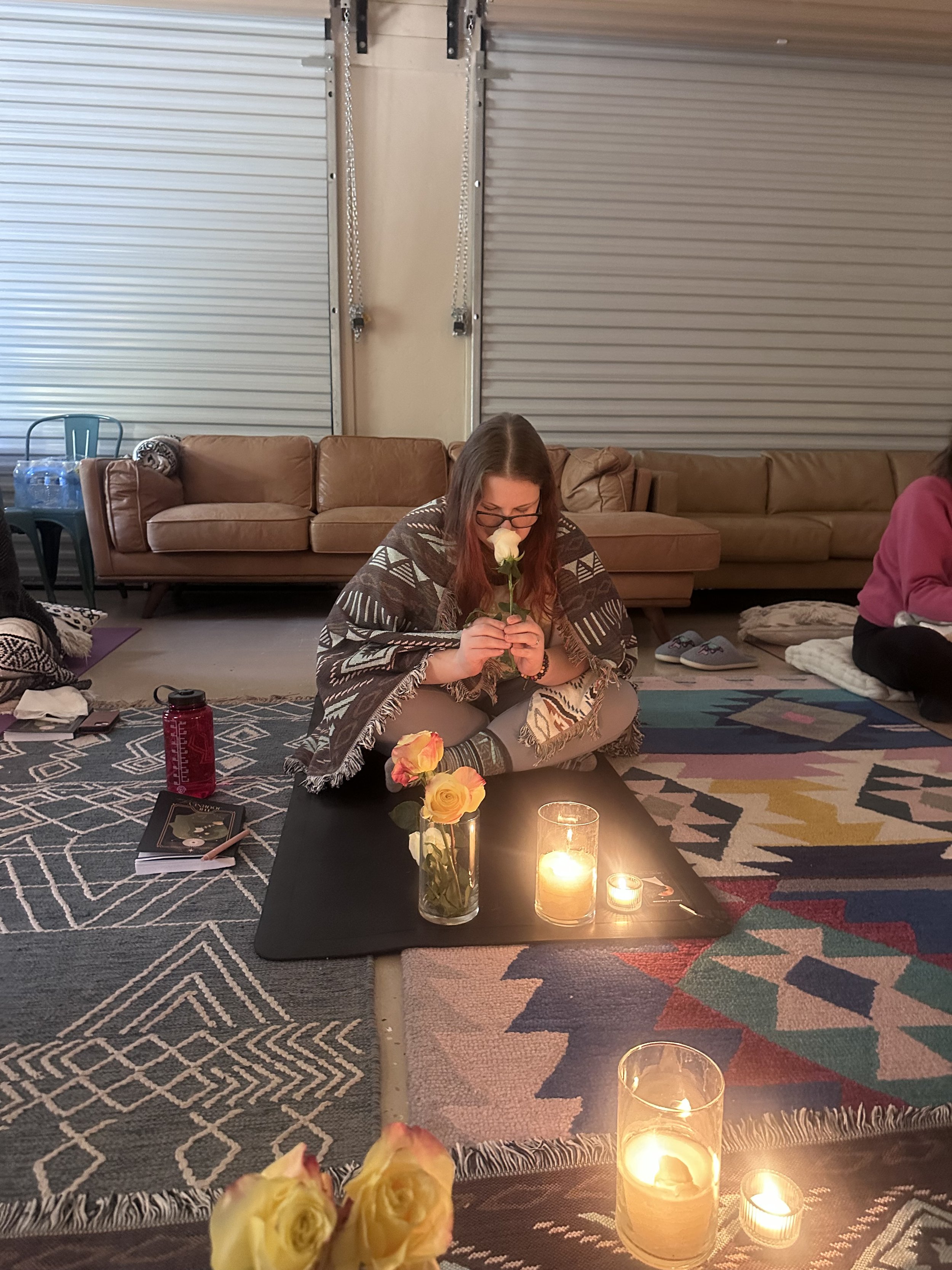 A woman sitting cross-legged on a rug holding a white rose near her face, surrounded by candles and flowers, in a cozy indoor setting with couches and blankets.