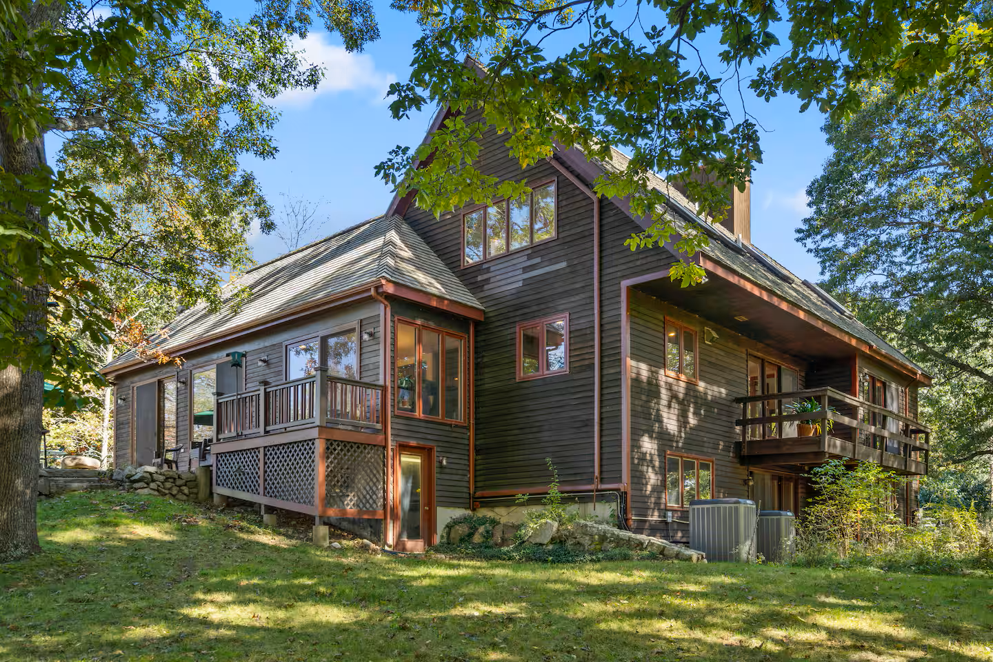 A large dark wooden house with multiple balconies and windows, situated in a wooded area with green grass and trees.