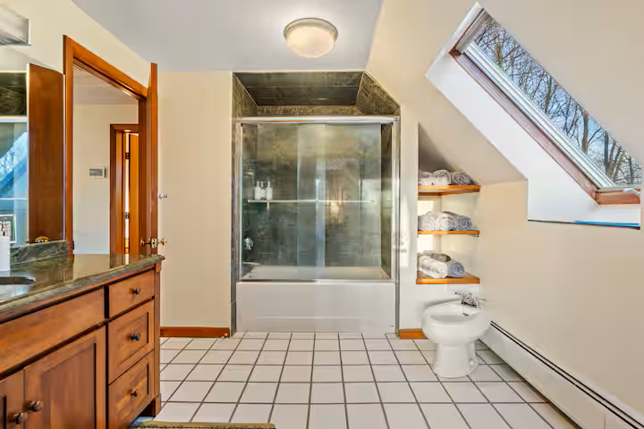 Bathroom with a shower enclosure, a toilet, wooden shelves with towels, a skylight window, and a wooden cabinet.