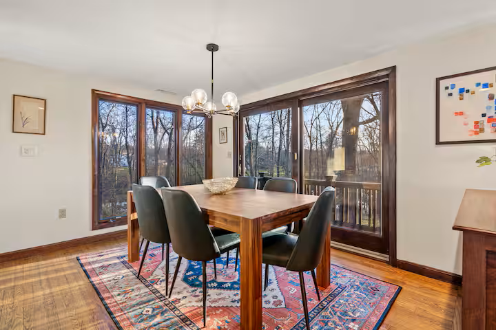 Dining room with a wooden table, six black chairs, a colorful patterned rug, large windows with wooden frames, a modern chandelier, and framed artwork on the walls.
