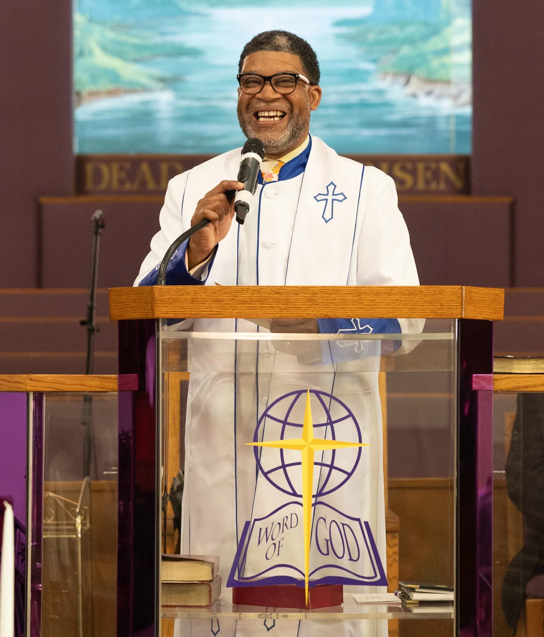 A man in a white clergy robe with blue and yellow accents, holding a microphone and speaking at a church podium. The podium displays a Christian cross, a globe, and the words 'Word of God.' The man is smiling with glasses, in front of a church interior background.