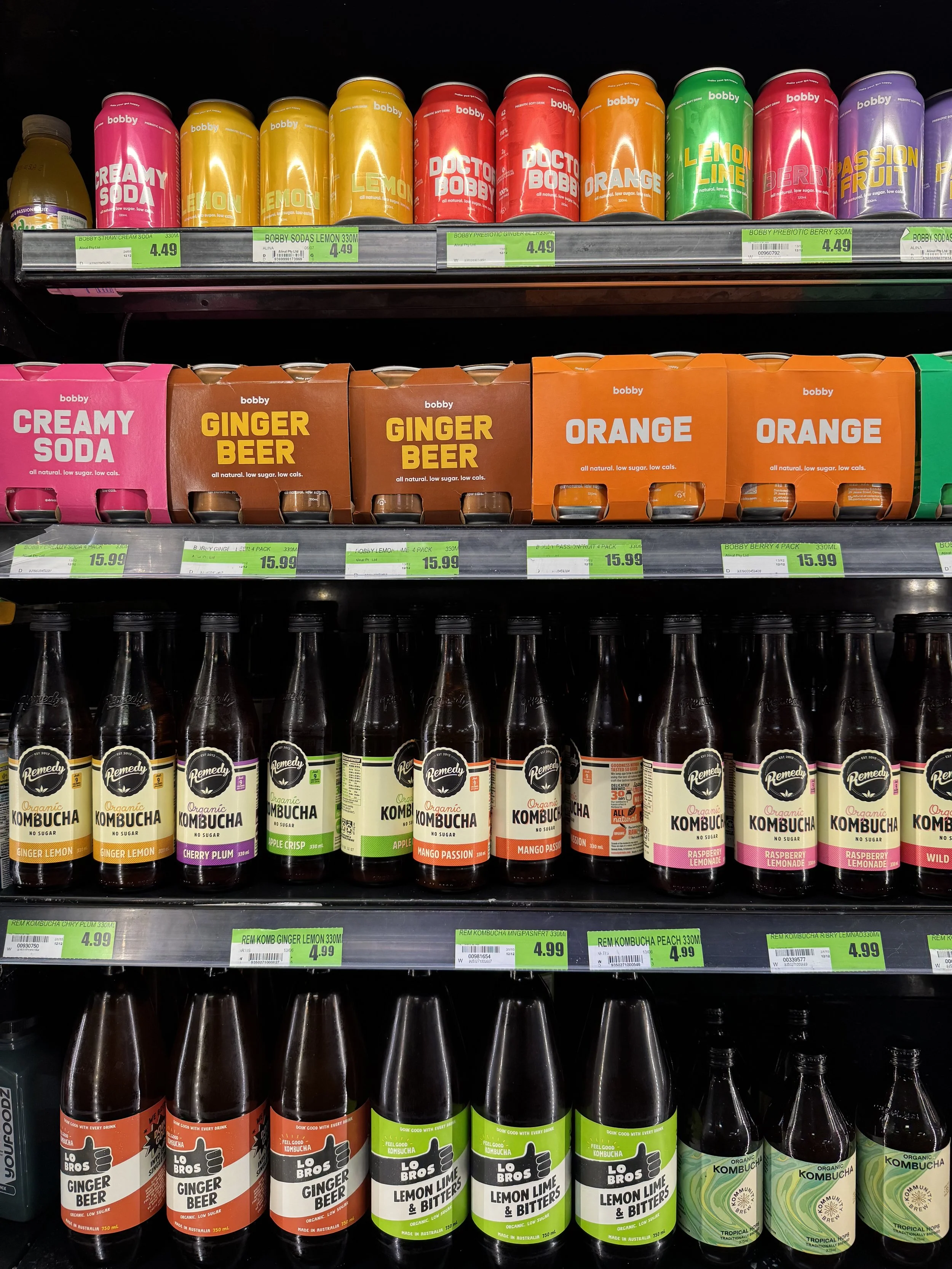 A grocery store shelf displaying various canned sodas, boxed ginger beers, and bottles of kombucha in different flavors.