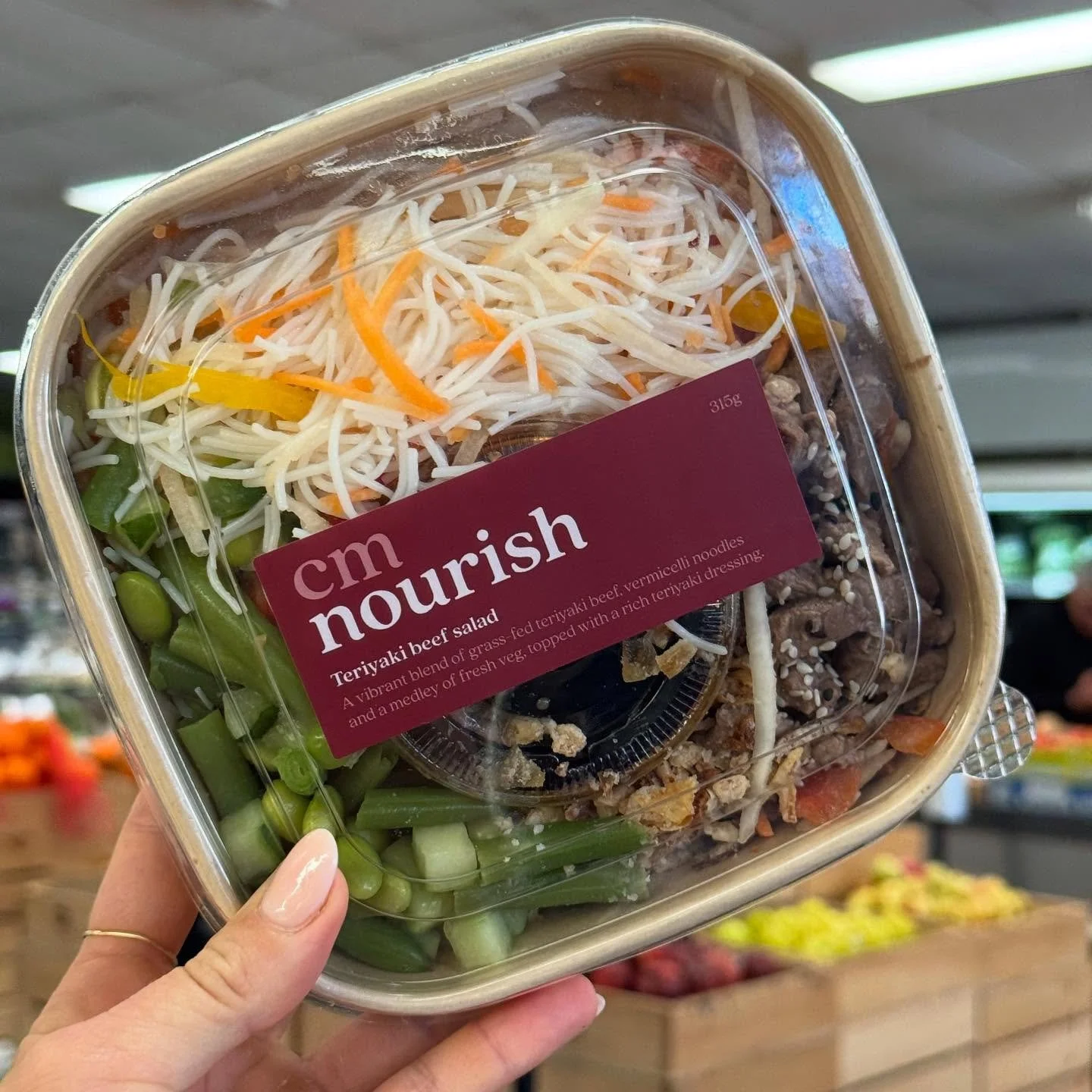 A hand holding a plastic container with a terryaki beef salad inside. The salad includes shredded vegetables, green beans, and beef with sesame seeds. The container has a purple label that reads "cm nourish, terryaki beef salad."