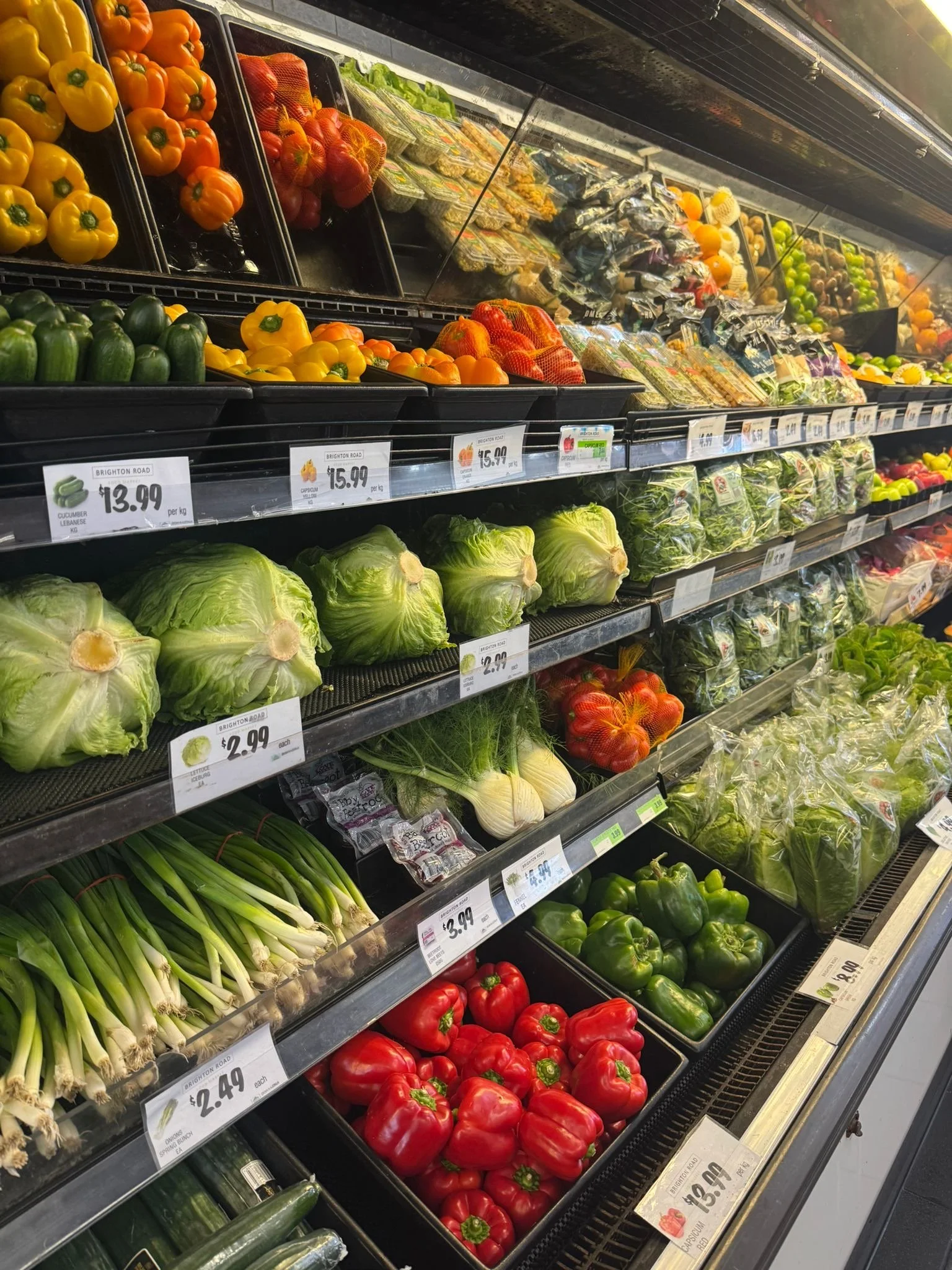 Fresh vegetables including yellow, orange, and green bell peppers, lettuce, tomatoes, cucumbers, green onions, and other produce in a grocery store refrigerated display case.