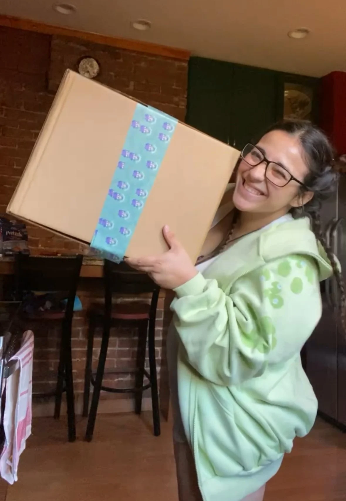 Woman smiling and holding a large wrapped gift box in a cozy indoor setting.