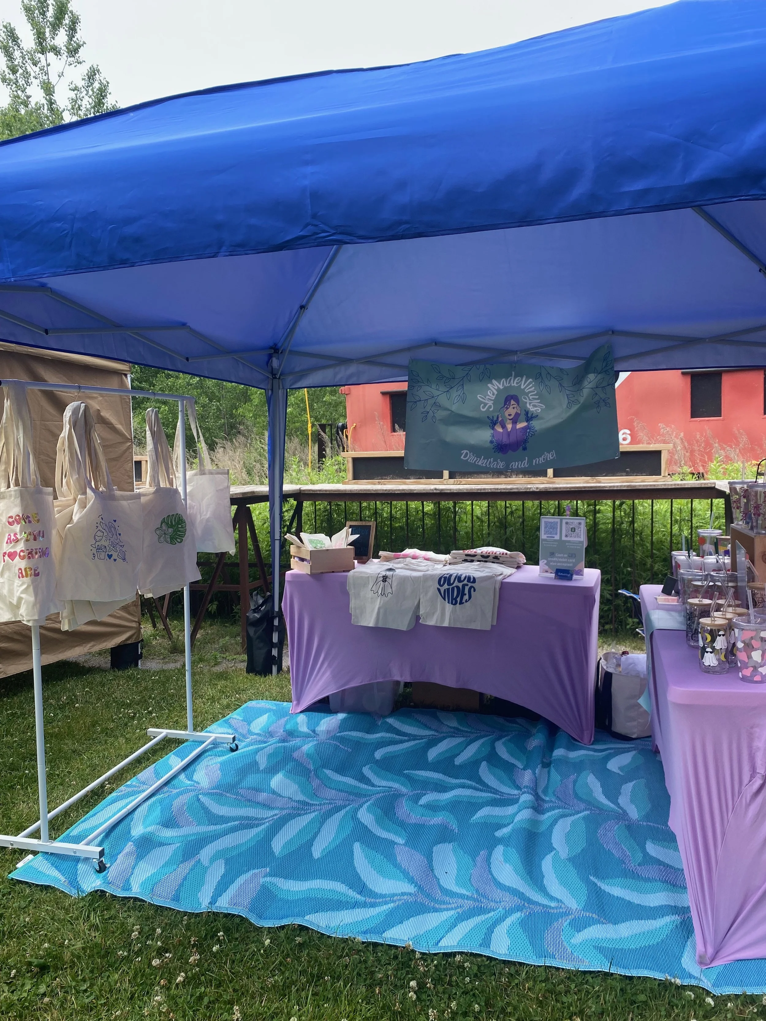 An outdoor booth at a market with tote bags and glassware under a blue canopy, on a grassy area with a pink building and trees in the background.