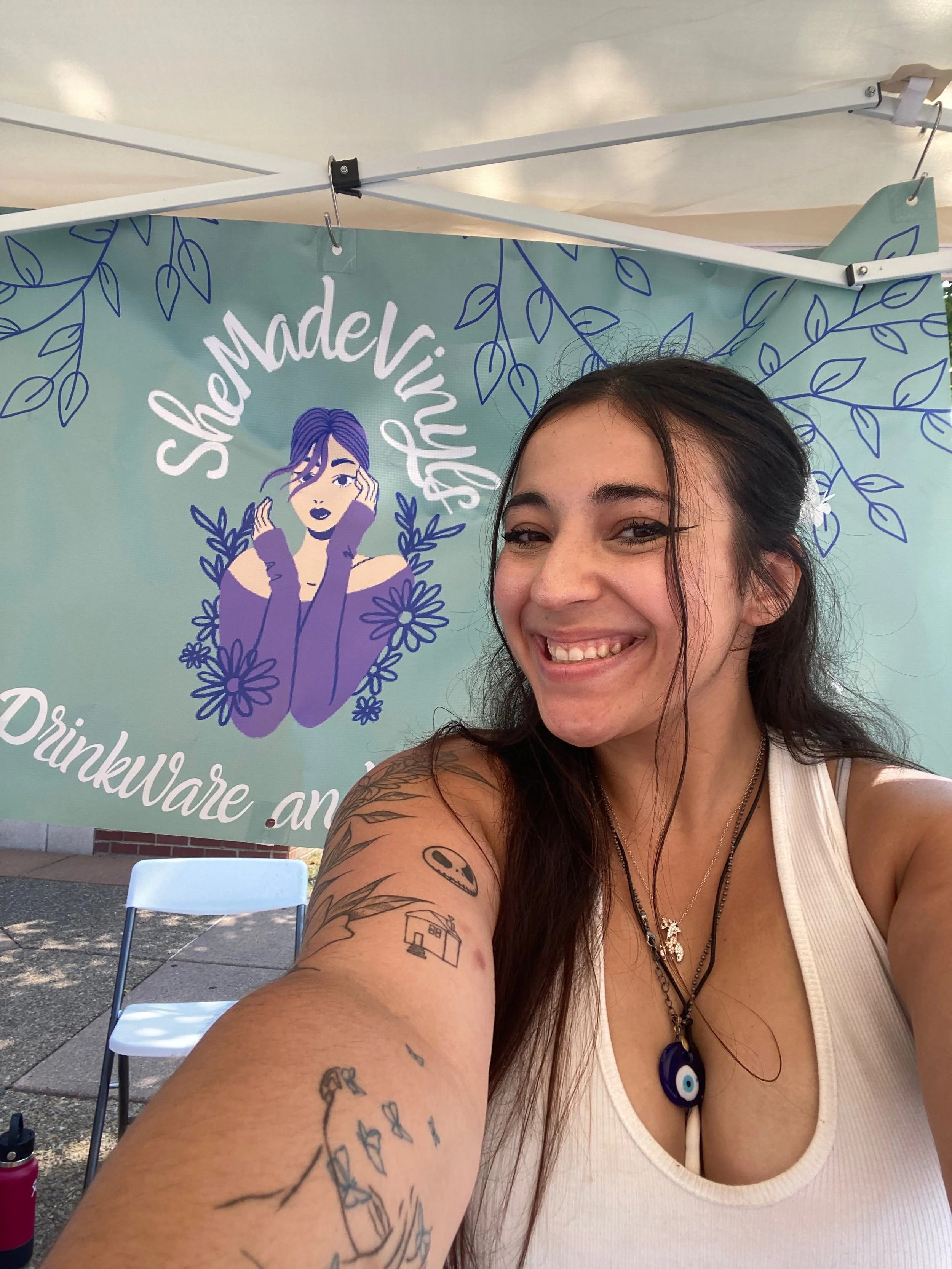 Young woman smiling and taking a selfie at a booth with a banner behind her that reads "SheMadeVinyls" with an illustration of a woman and floral decorations. She has tattoos on her arm and is wearing necklaces, including a blue evil eye pendant.