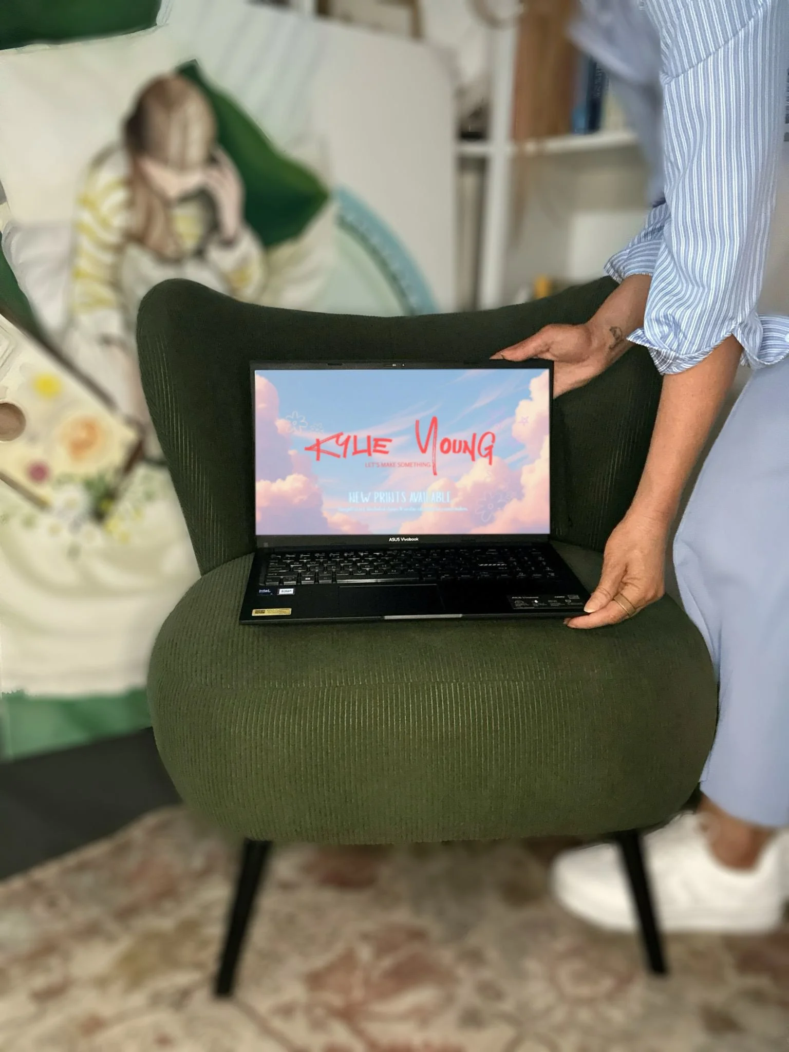 A person in a blue and white striped shirt and light-colored pants holds a black ASUS laptop with a colorful cloudy sky background and red text on the screen that reads "Kyke Young." The person is standing next to a green upholstered chair, and a wom