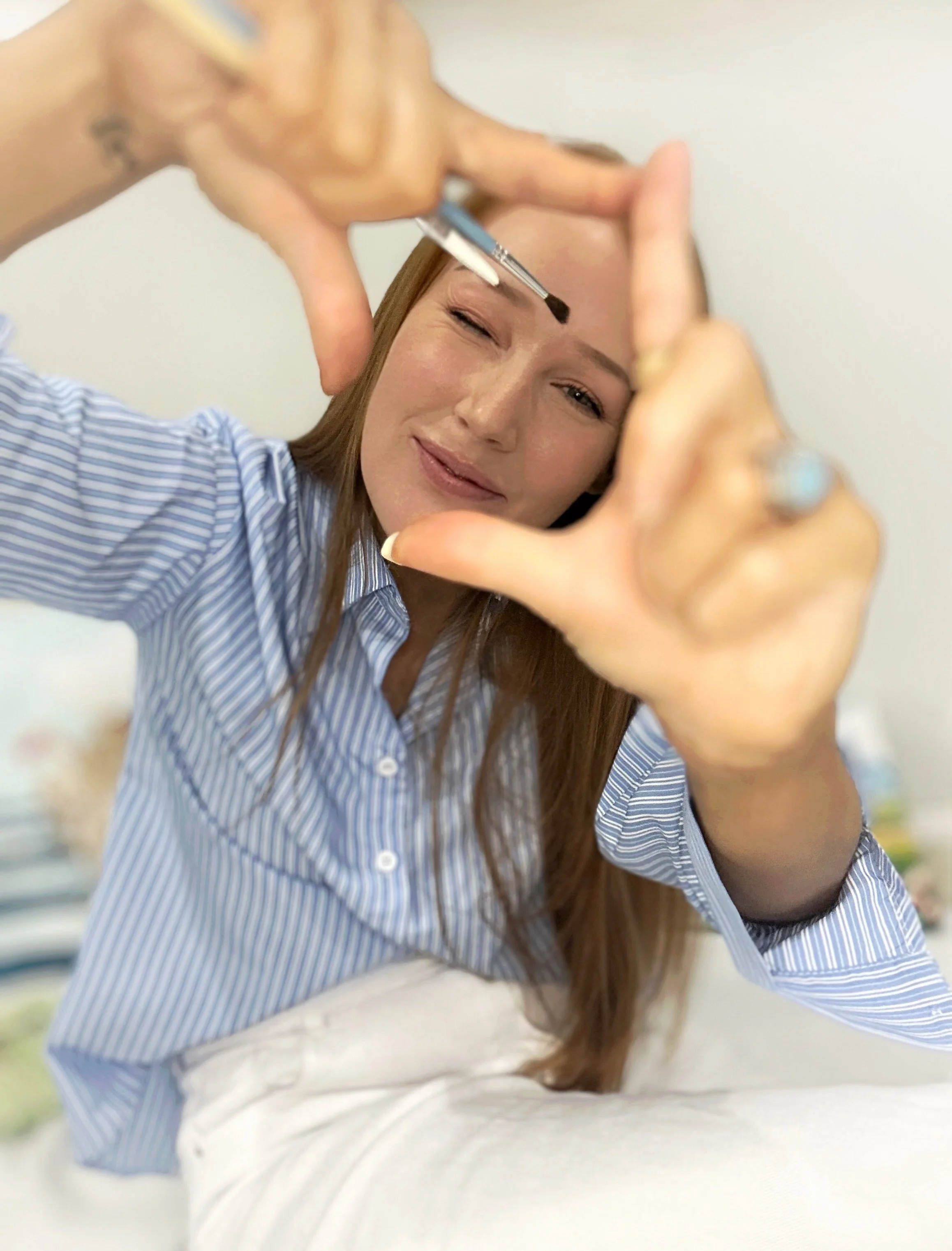 Woman with long brown hair in a blue striped shirt making a frame with her hands, holding a small makeup brush near her face, smiling slightly while winking.