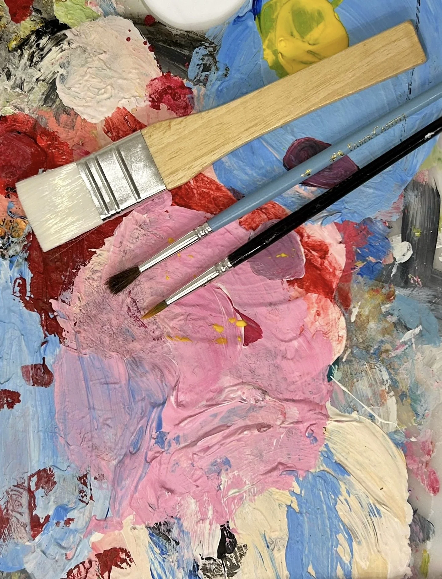 Paint palette with thick layers of pink, blue, red, yellow, white, and black acrylic paint, along with three paintbrushes resting on it.