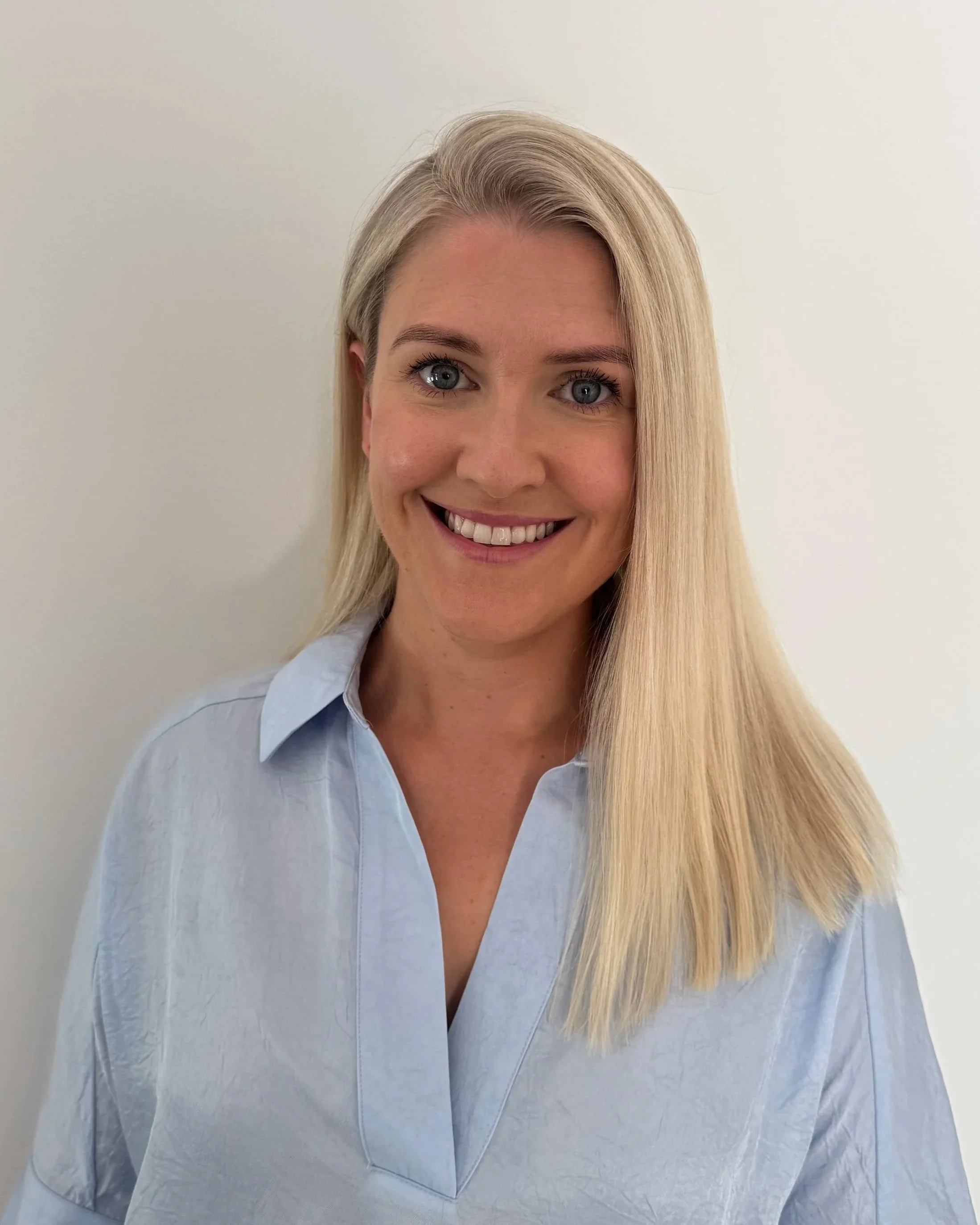 Close-up of a smiling woman with blonde hair wearing a light blue button-up shirt against a plain white background.