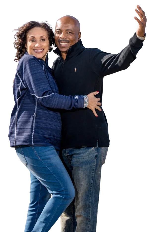 A smiling middle-aged couple taking a selfie outdoors, with the woman holding the camera, both wearing casual dark-colored clothing and jeans.