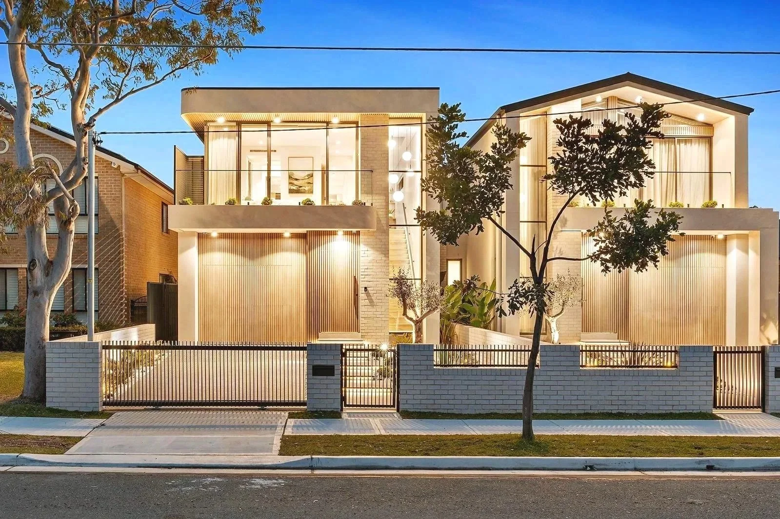 Modern two-story house with large glass windows illuminated at dusk, surrounded by a white brick fence, small trees, and a sidewalk.