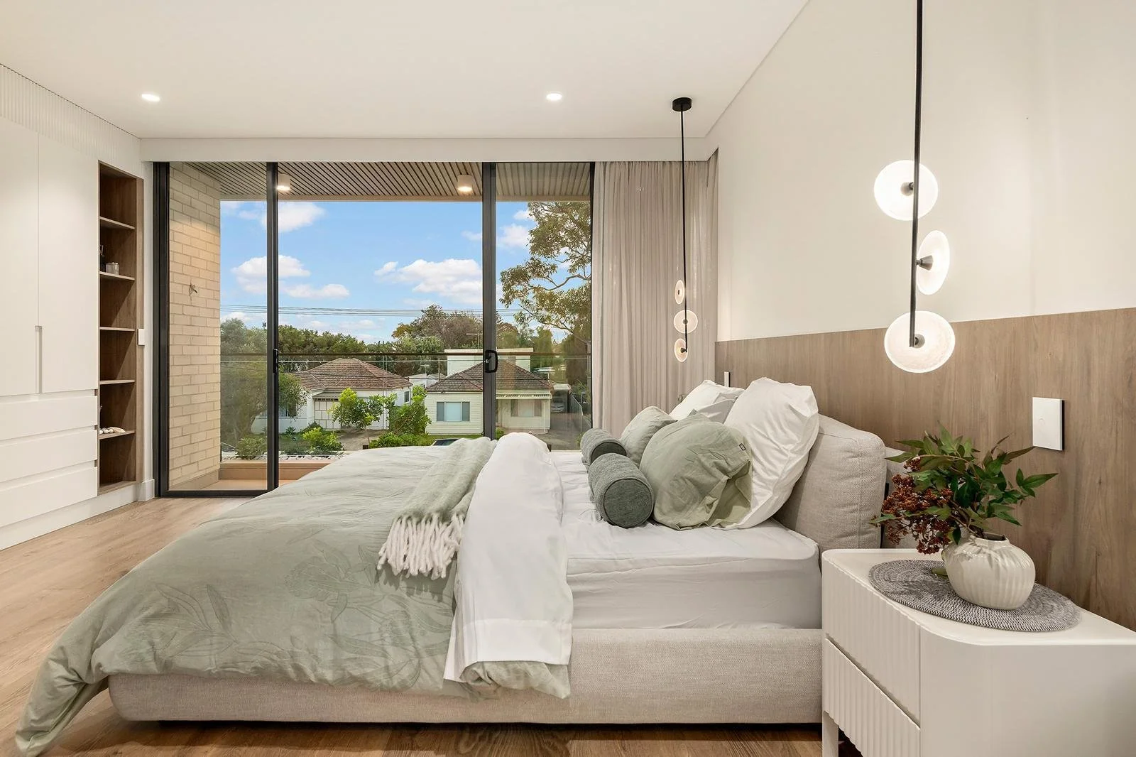 Modern bedroom with a large bed, bedside table with a plant, and hanging pendant lights. Floor-to-ceiling windows showing a view of houses and trees outside.