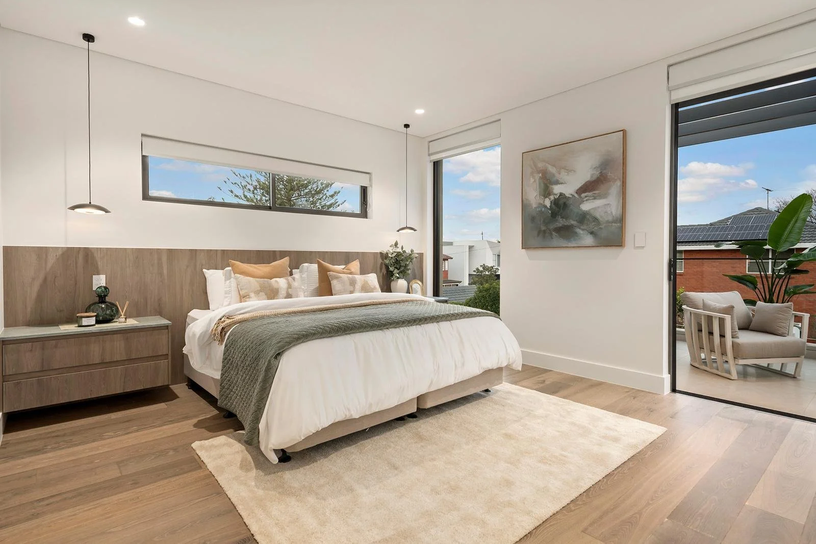Modern bedroom with white walls, a large bed with neutral linens and pillows, a wooden nightstand with decorative items, wall-mounted pendant lights, and a balcony with outdoor seating and solar panels visible on neighboring rooftops.