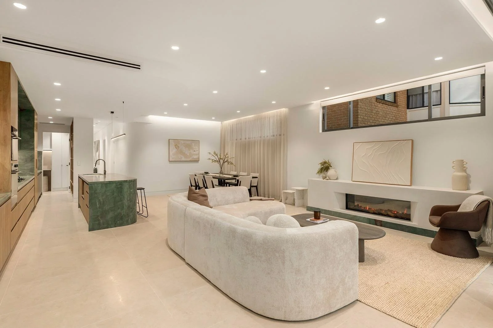Modern living room with a white curved sofa, fireplace, and brown and beige accents, open to a dining area and kitchen.