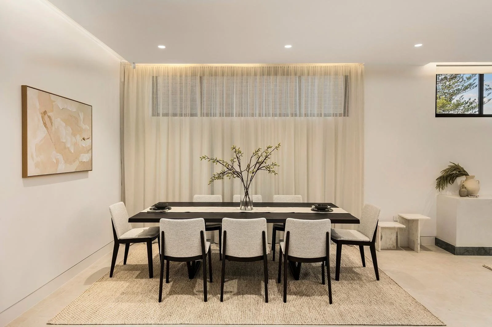 Minimalist dining room with a dark wood table, eight light-colored chairs, a large glass vase with branches, and neutral decor.