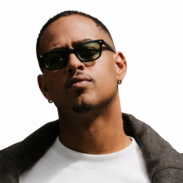Portrait of a young man wearing sunglasses, has small earrings, a goatee, and is dressed in a white shirt with a dark jacket over his shoulder.
