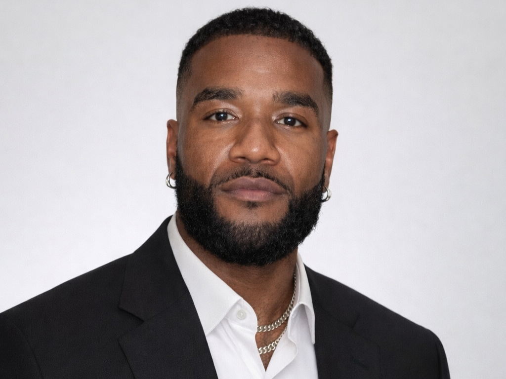 A professional headshot of a Black man with a beard, short curly hair, wearing a black suit jacket, white shirt, and silver jewelry, against a plain white background.