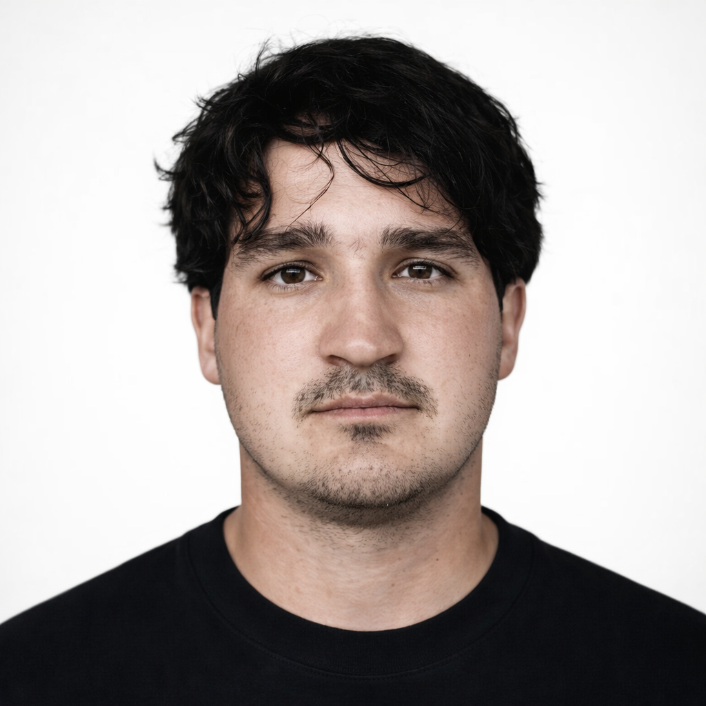 Close-up portrait of a young man with dark, wavy hair, light skin, and brown eyes, wearing a black shirt against a plain white background.