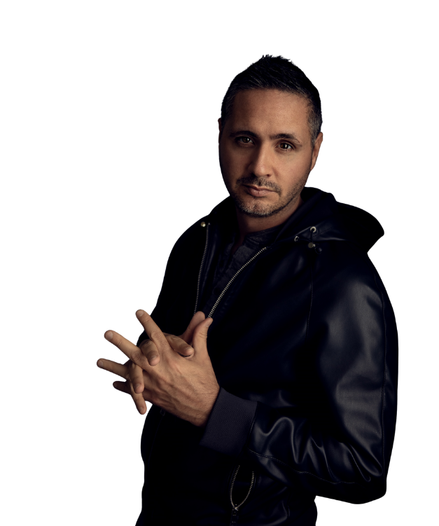 A man with short dark hair and a slight beard, wearing a black leather jacket, posing with hands clasped in front of a black background.
