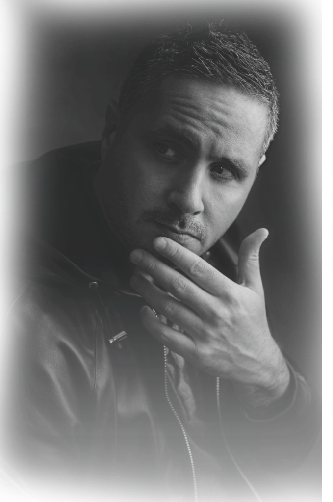 Black and white portrait of a man with short hair, wearing a leather jacket, looking thoughtfully to the side with his hand near his chin.