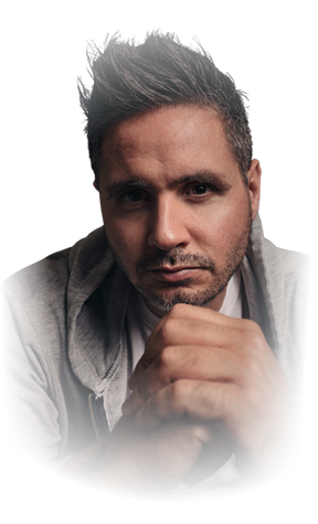 A man with dark, spiked hair and light facial hair is looking directly at the camera. He is resting his chin on folded hands against a black background.