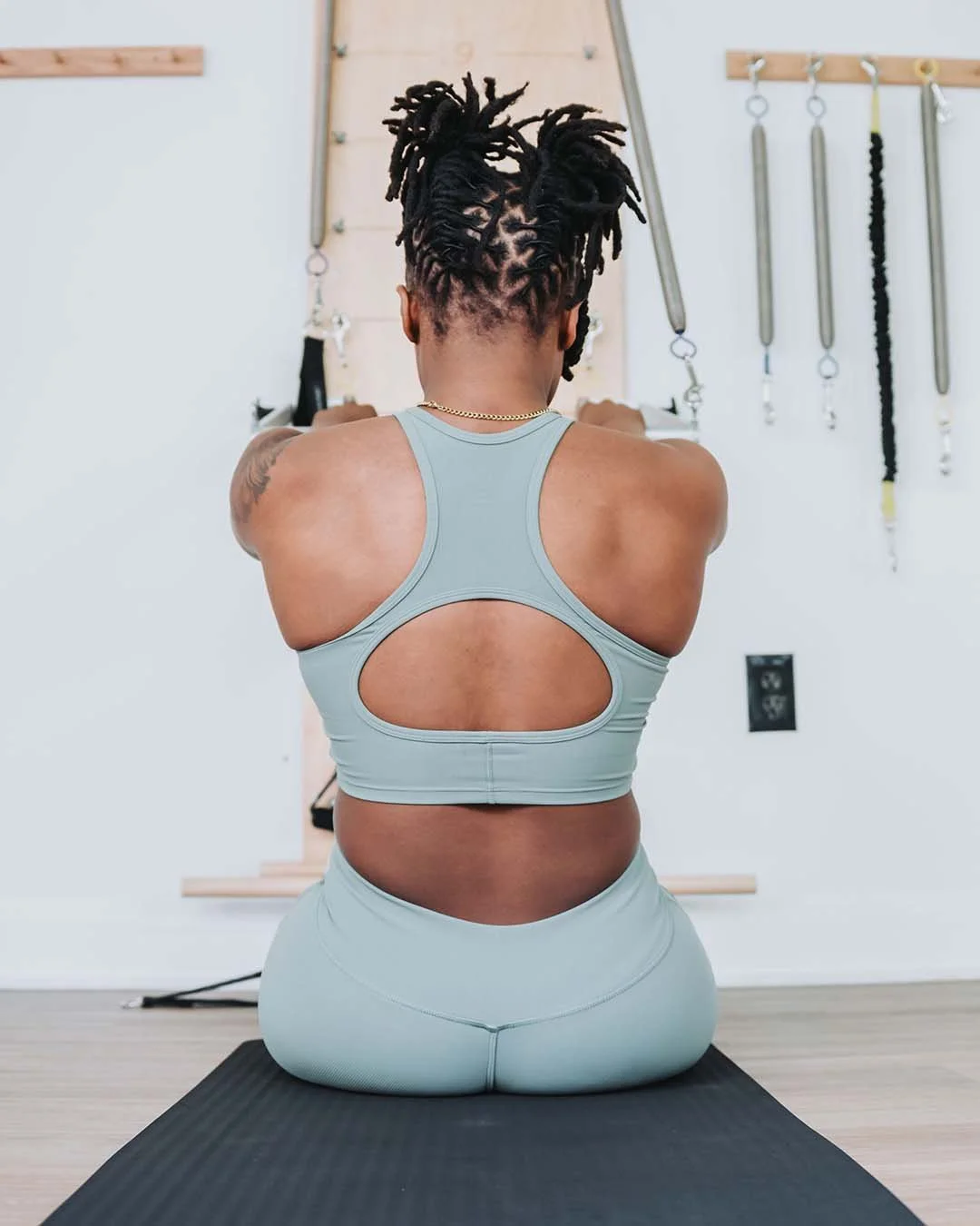 Back view of a woman doing yoga in a light gray sports bra and leggings, kneeling on a yoga mat in a fitness studio with hanging resistance bands on the wall.