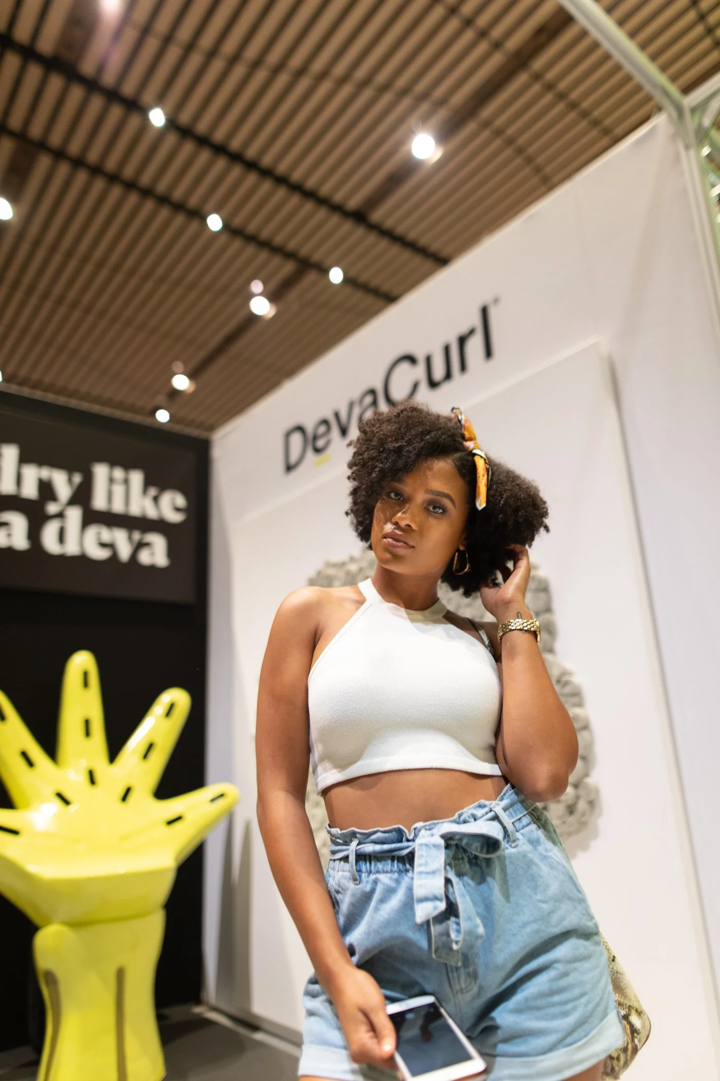 A woman with curly hair, wearing a white crop top and denim shorts, standing in an indoor art or design exhibit, with a yellow hand-shaped sculpture behind her and signage that reads "DevraGurl" and some text in the background.