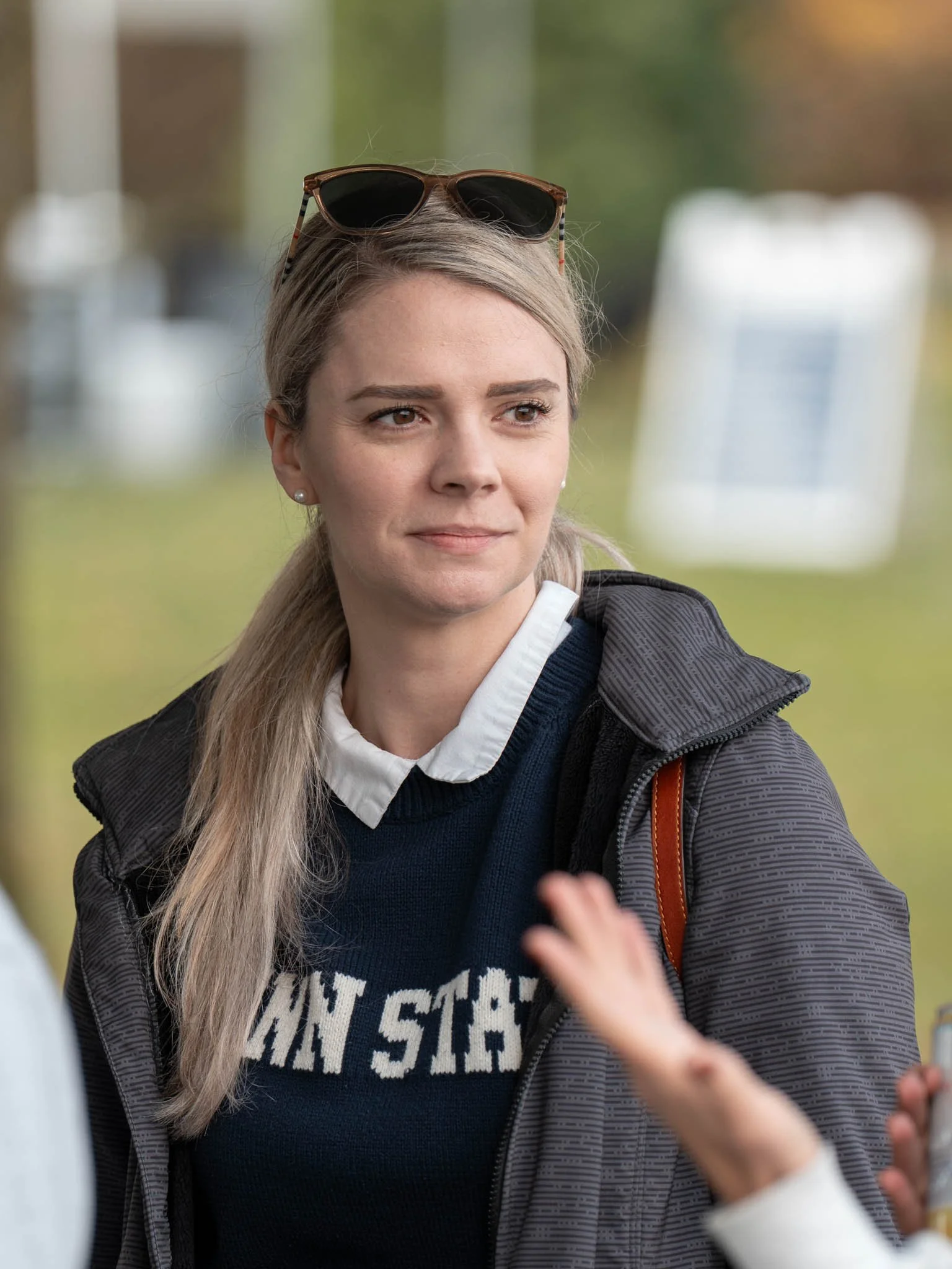 A woman with blonde hair wearing sunglasses on her head, a dark jacket, a white collar shirt, and a navy sweater with white across the chest, is outdoors. She appears to be listening to someone, with a serious or neutral expression on her face.