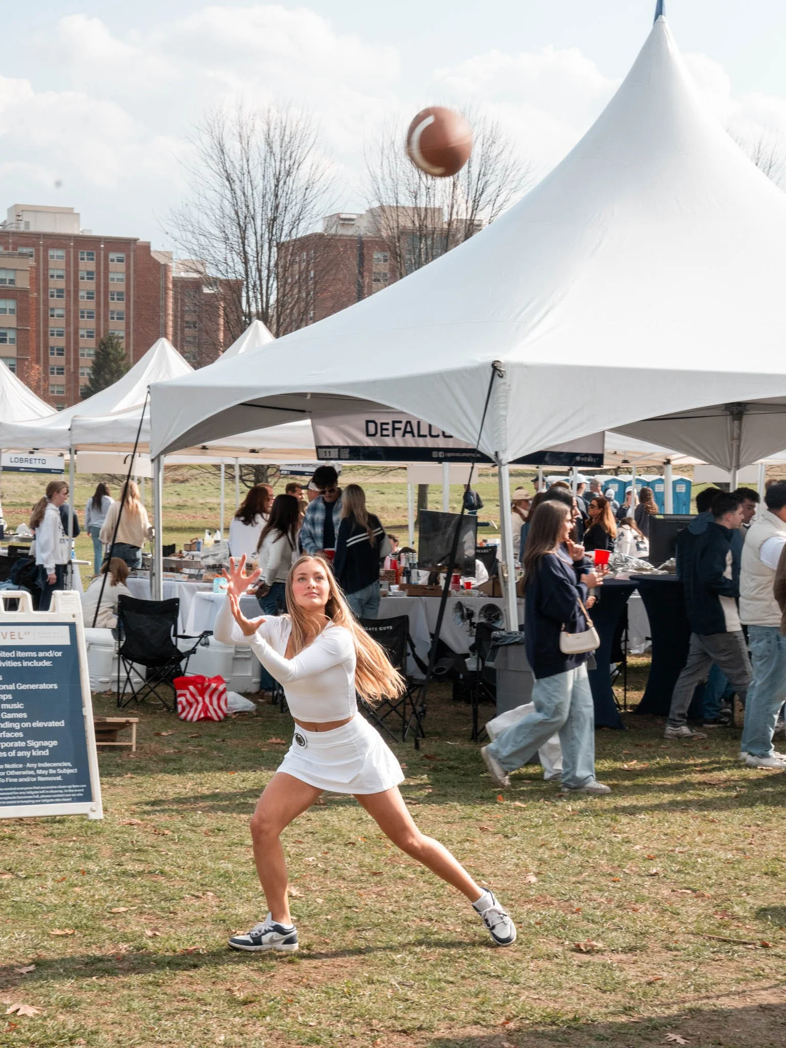 A young woman in athletic clothes throws a football at an outdoor event with tents and people in the background.