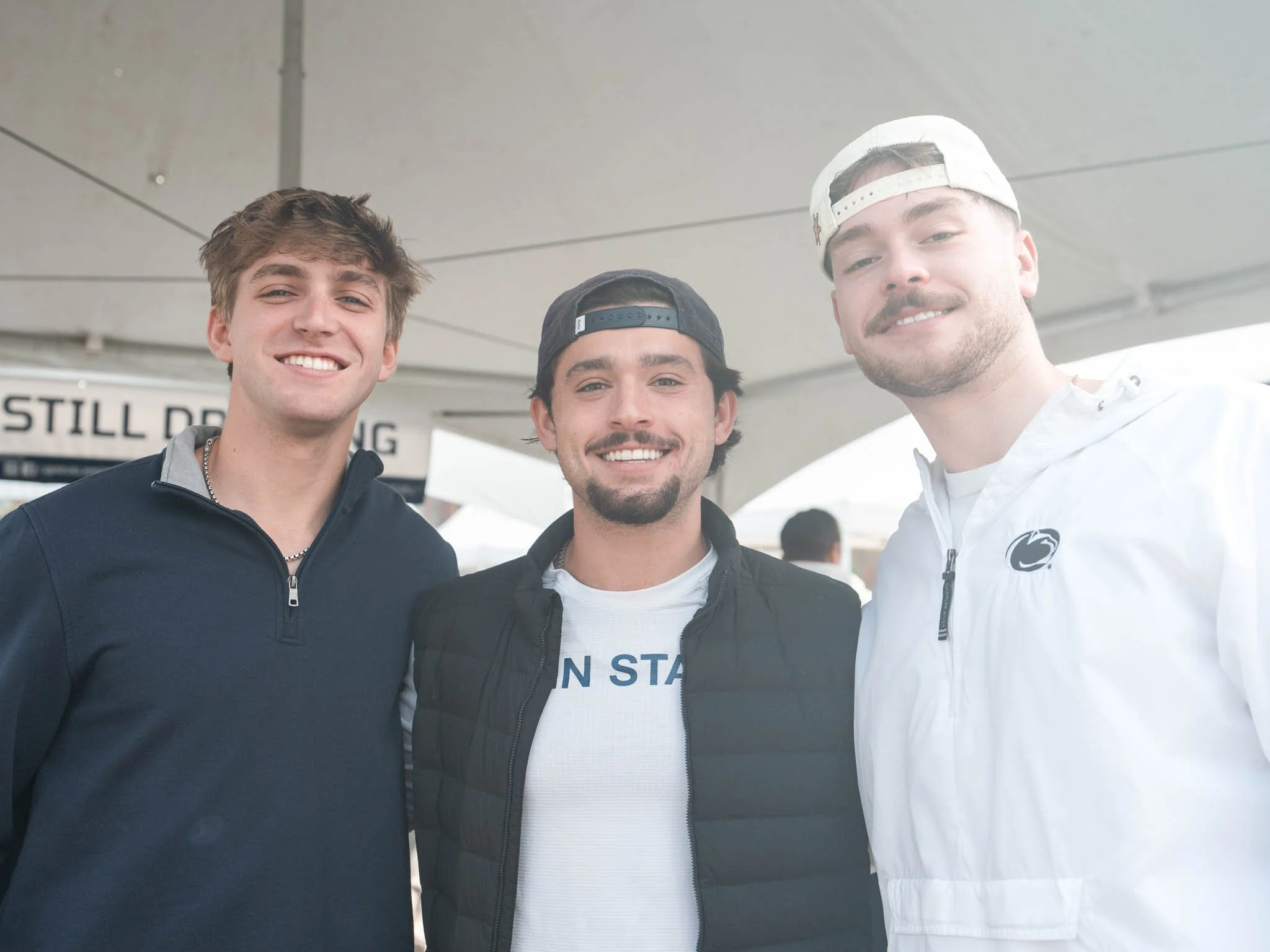 Three young men standing close together, smiling at the camera under a large white canopy.