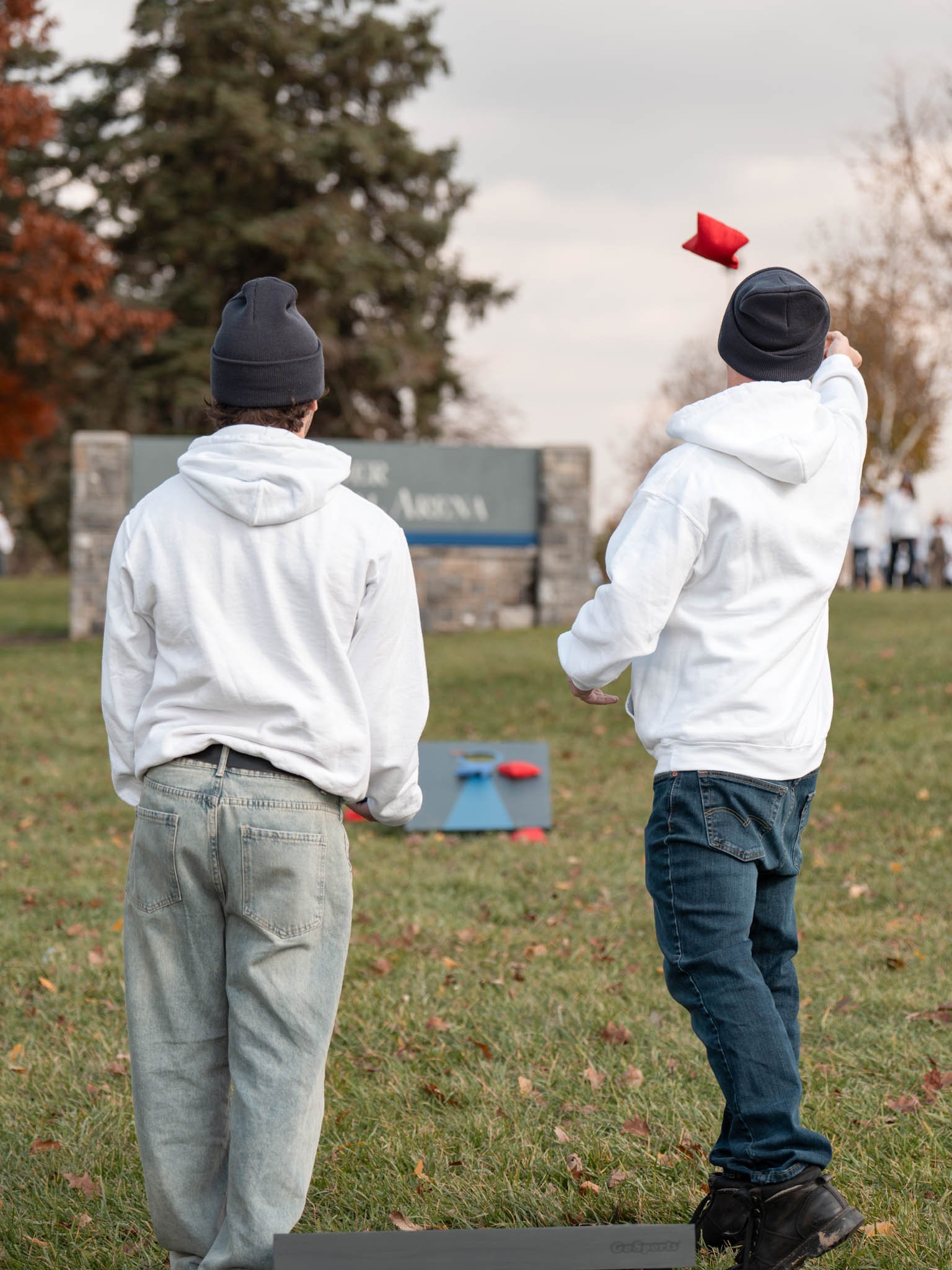 Two boys wearing white hoodies and black beanies playing a game of cornhole outdoors on a grassy field with trees and a sign in the background.