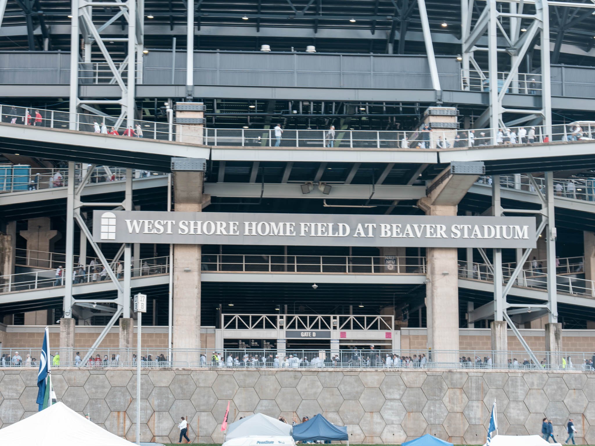 Exterior view of Beaver Stadium with a large sign reading 'West Shore Home Field at Beaver Stadium' and crowds of people walking around.