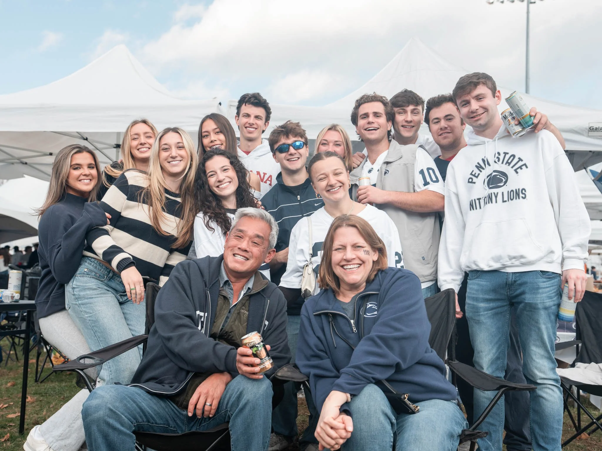 A group of smiling young adults and older adults posing together outdoors at a gathering or tailgate, with tents in the background.