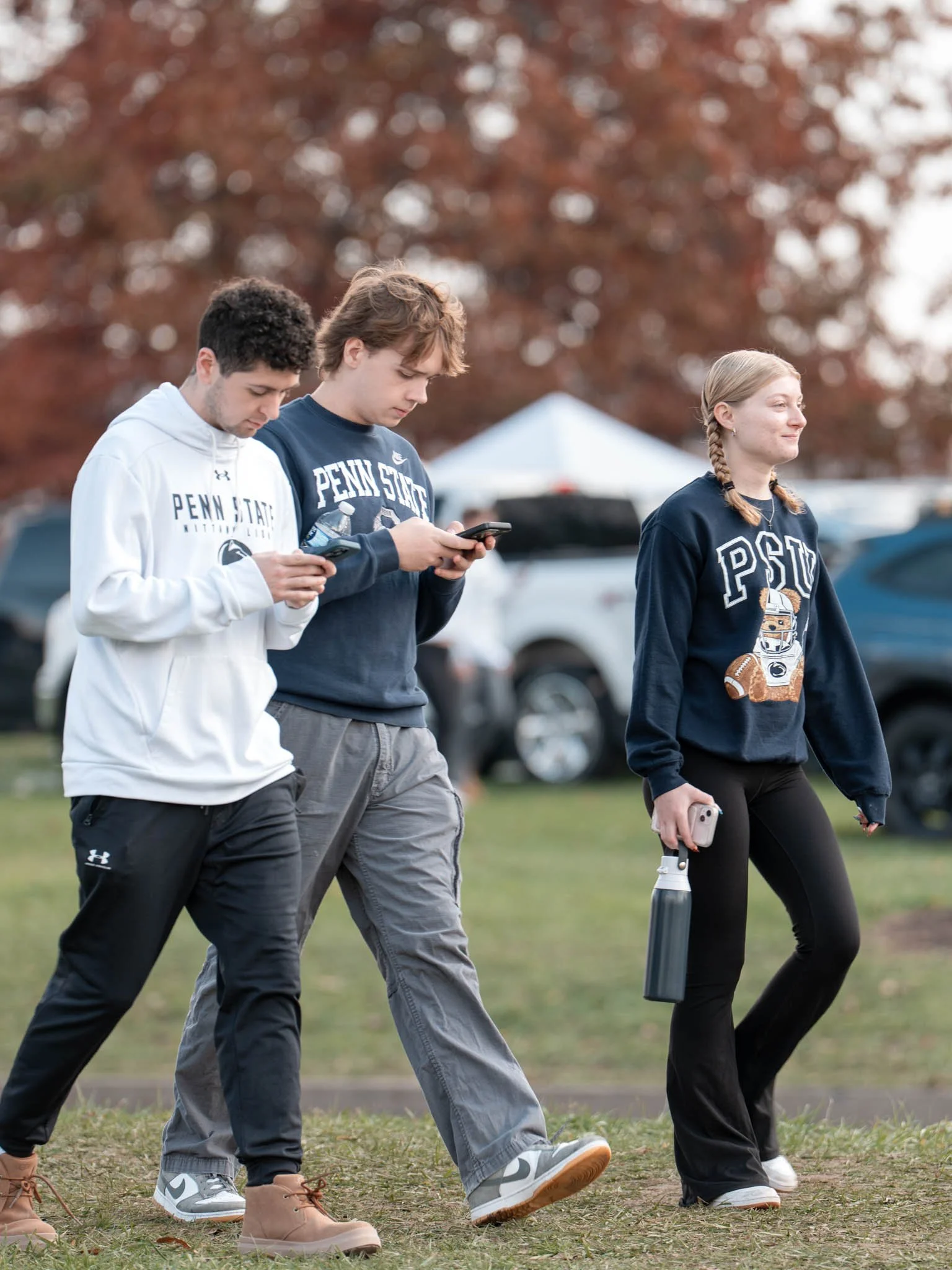 Three teenagers walking on grass, two are looking at smartphones and one is holding a water bottle and phone. They are wearing Penn State sweatshirts and casual pants. Background includes trees and cars.