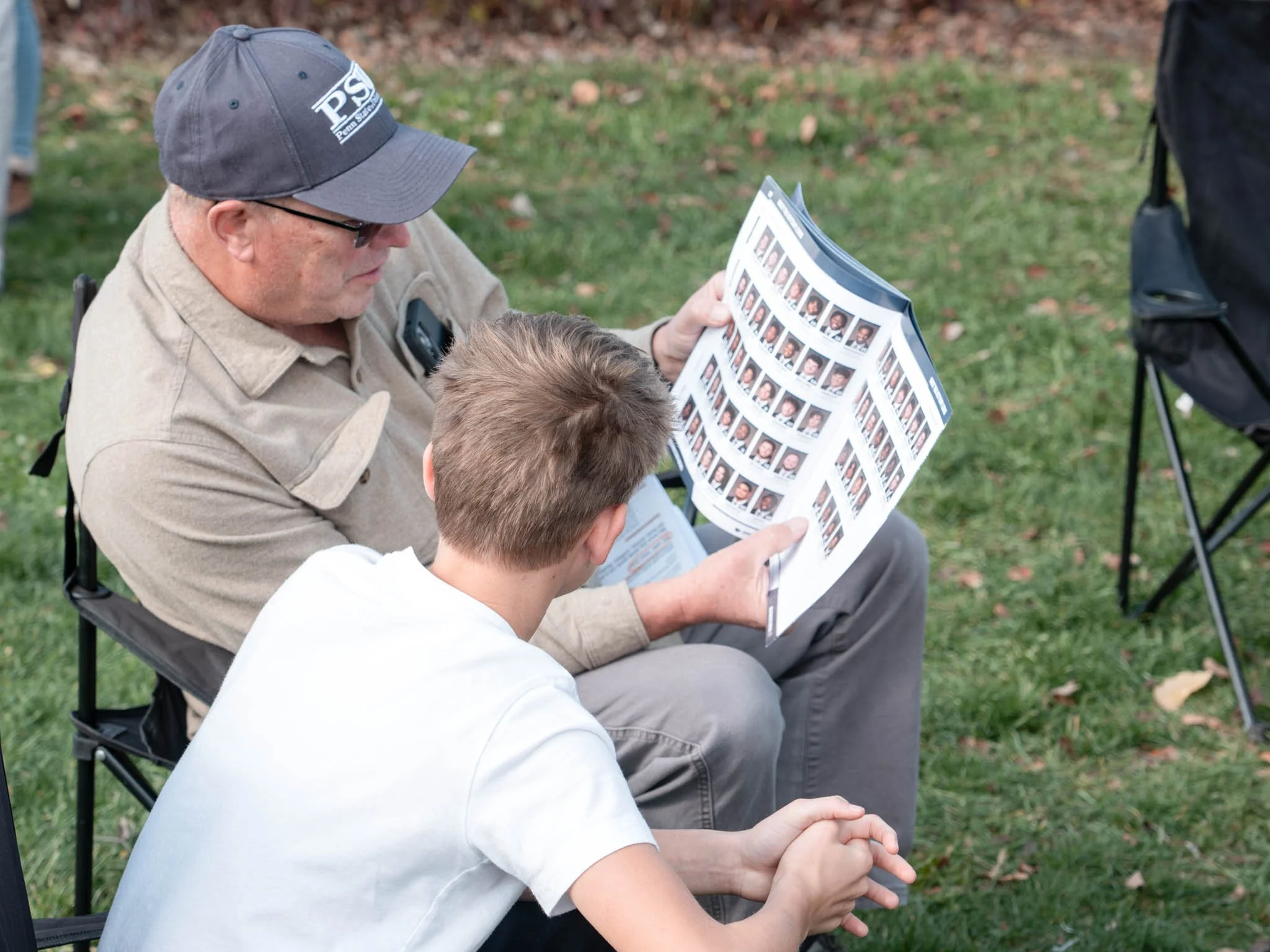 An older man and a young boy sitting outdoors on grass, looking at a sheet of photographs, possibly yearbook or school photos.