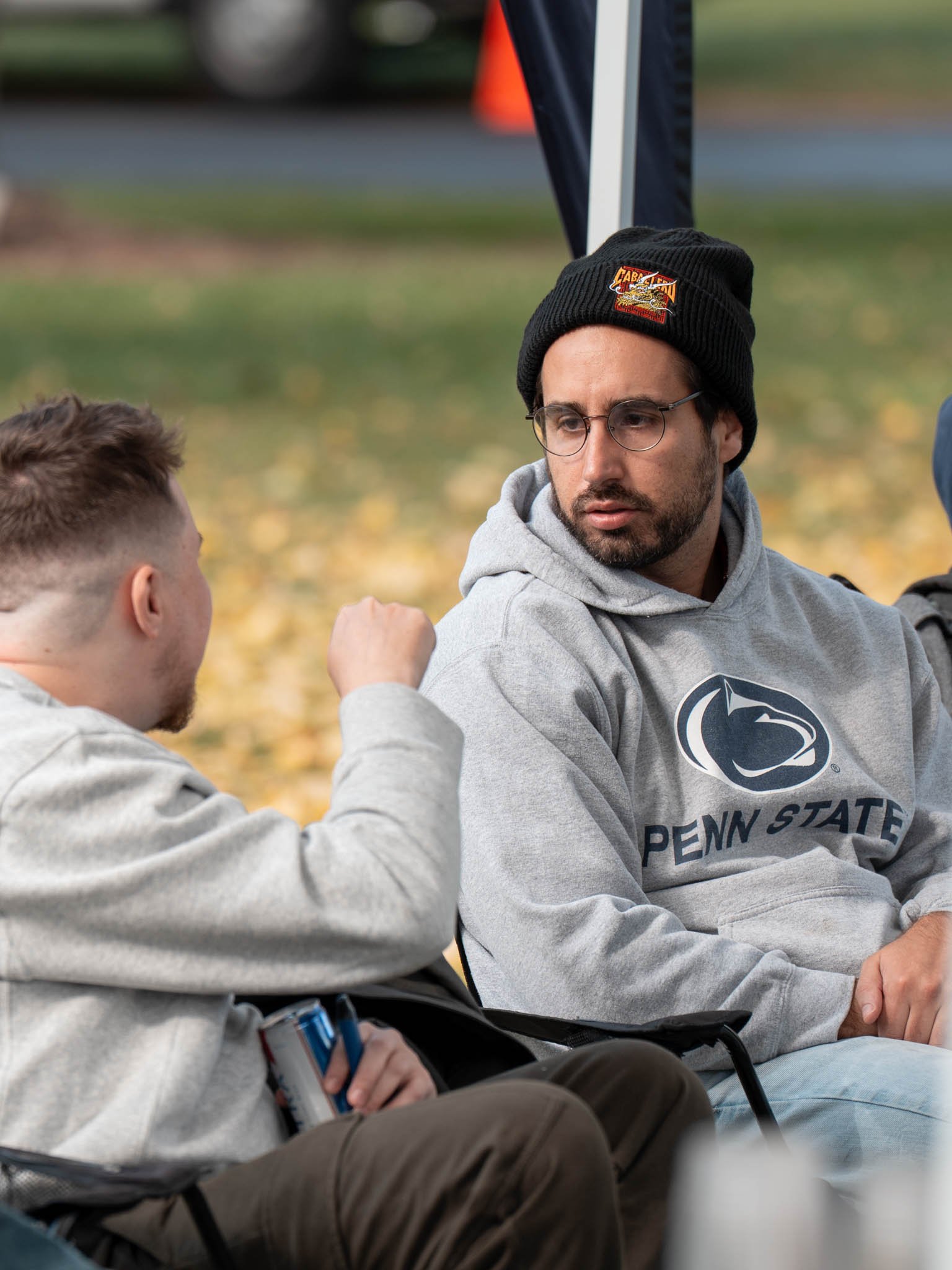 Two men sitting outdoors, one wearing a Penn State hoodie and a black beanie, the other in a light gray hoodie, engaged in conversation with autumn leaves on the ground.