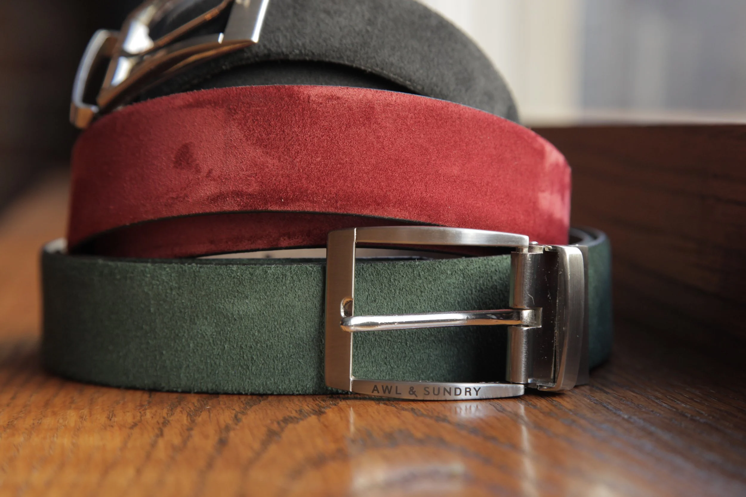 Stacked leather belts in black, red, and green with metal buckles, on a wooden surface.