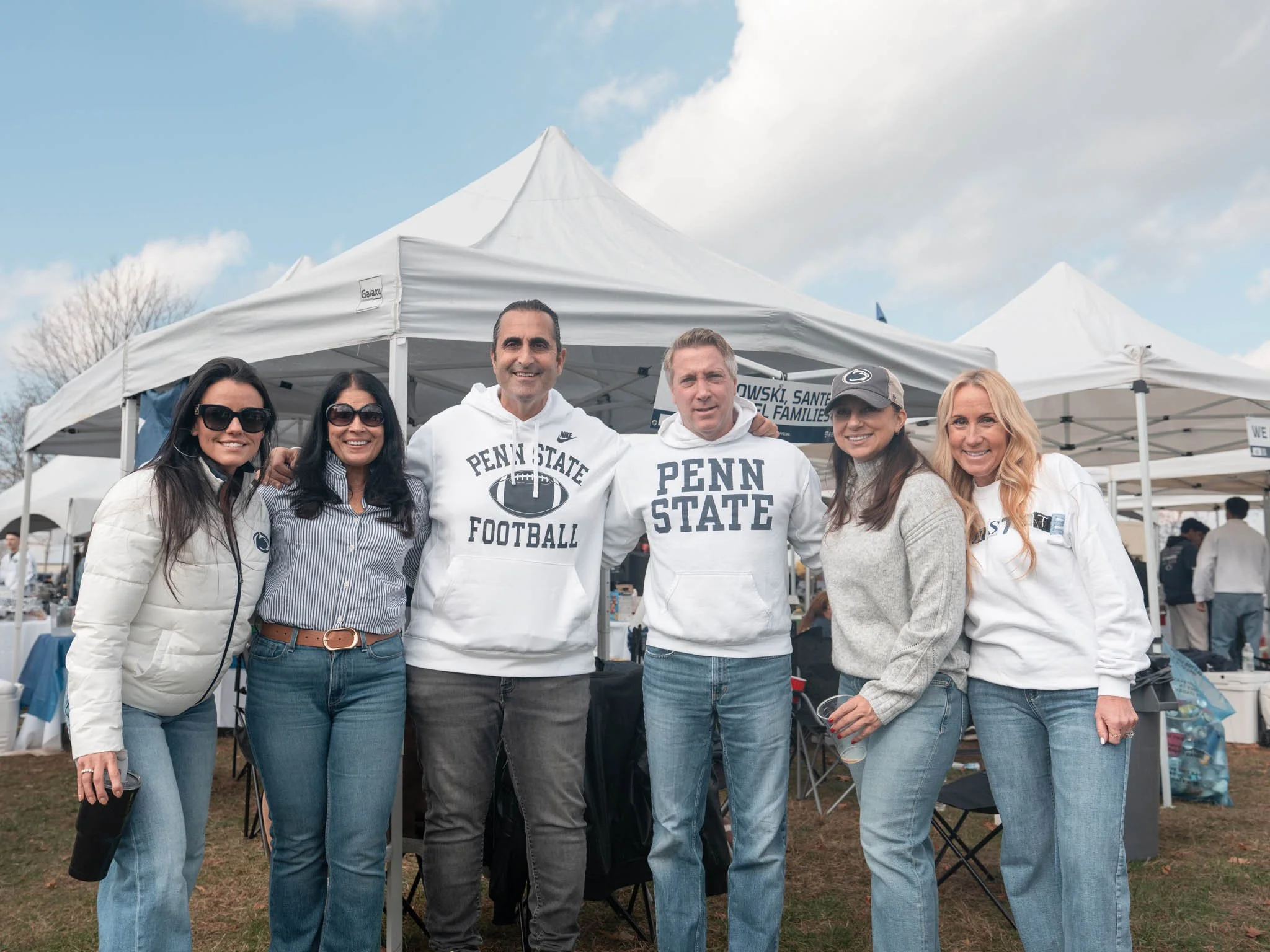Six people standing together outdoors in front of tents, smiling, wearing casual and Penn State apparel, at an outdoor event.