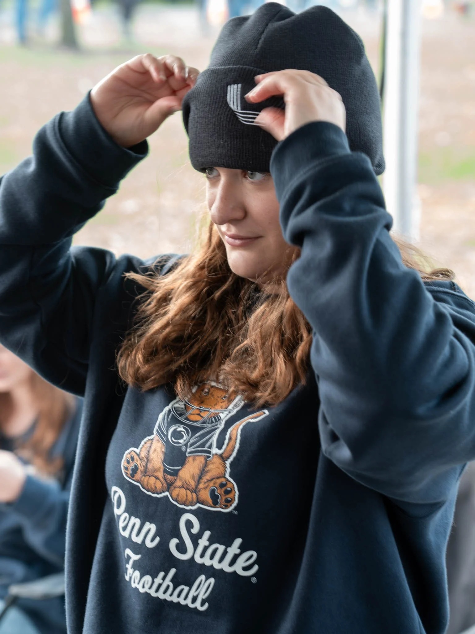 A woman with long red hair putting on a black beanie hat. She is wearing a Penn State football sweatshirt with a tiger mascot.