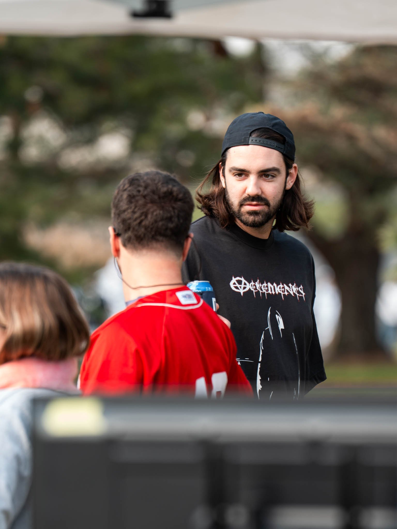 Three people outdoors, one man with long brown hair, beard, wearing a black cap backward and a black t-shirt with white text and graphic, talking to another person wearing a red sports jersey, with a man with glasses and a gray hoodie partially visib