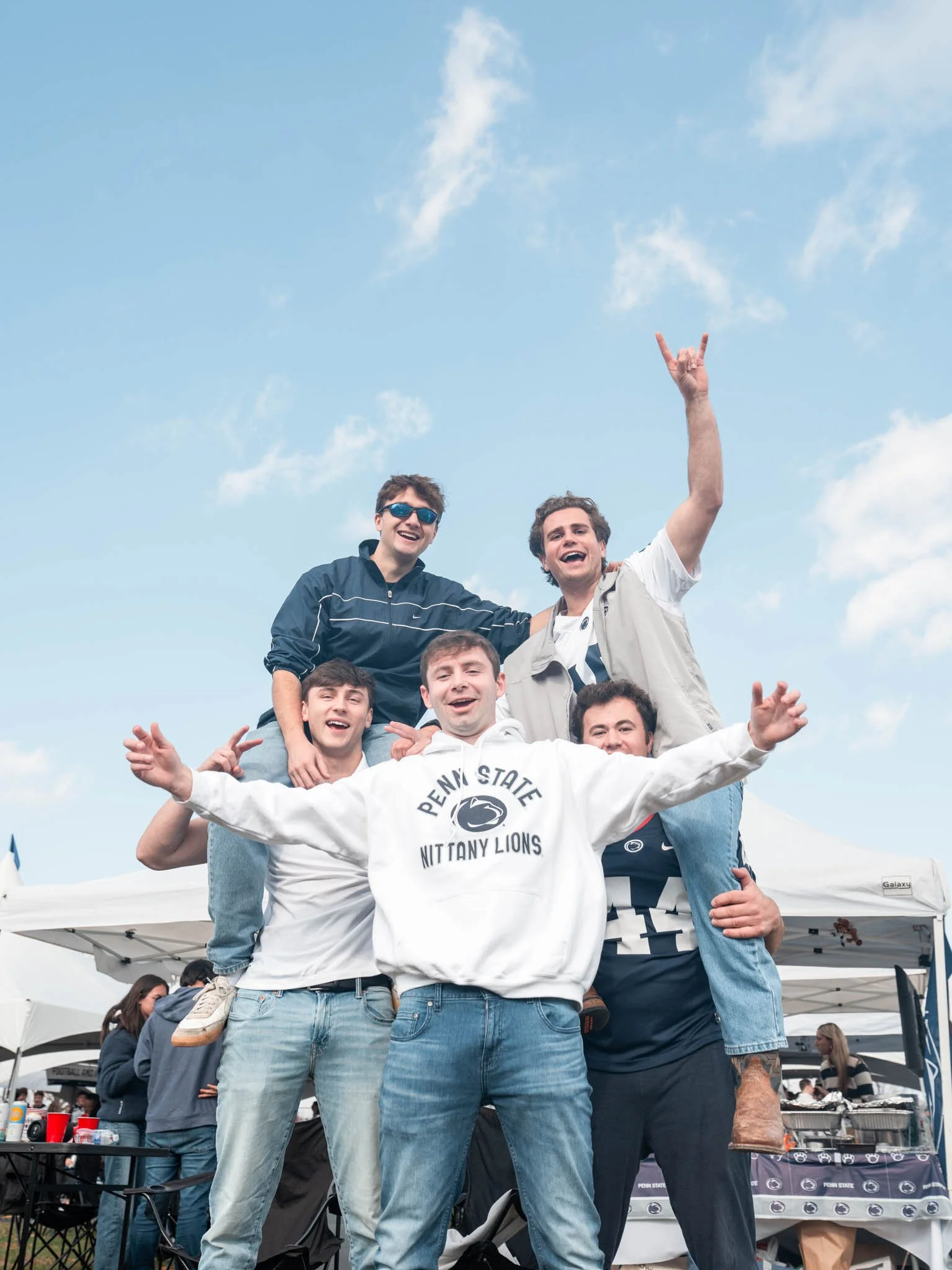 Group of five young men celebrating outdoors during a Penn State event, forming a human pyramid, with tents and other people in the background under a partly cloudy sky.