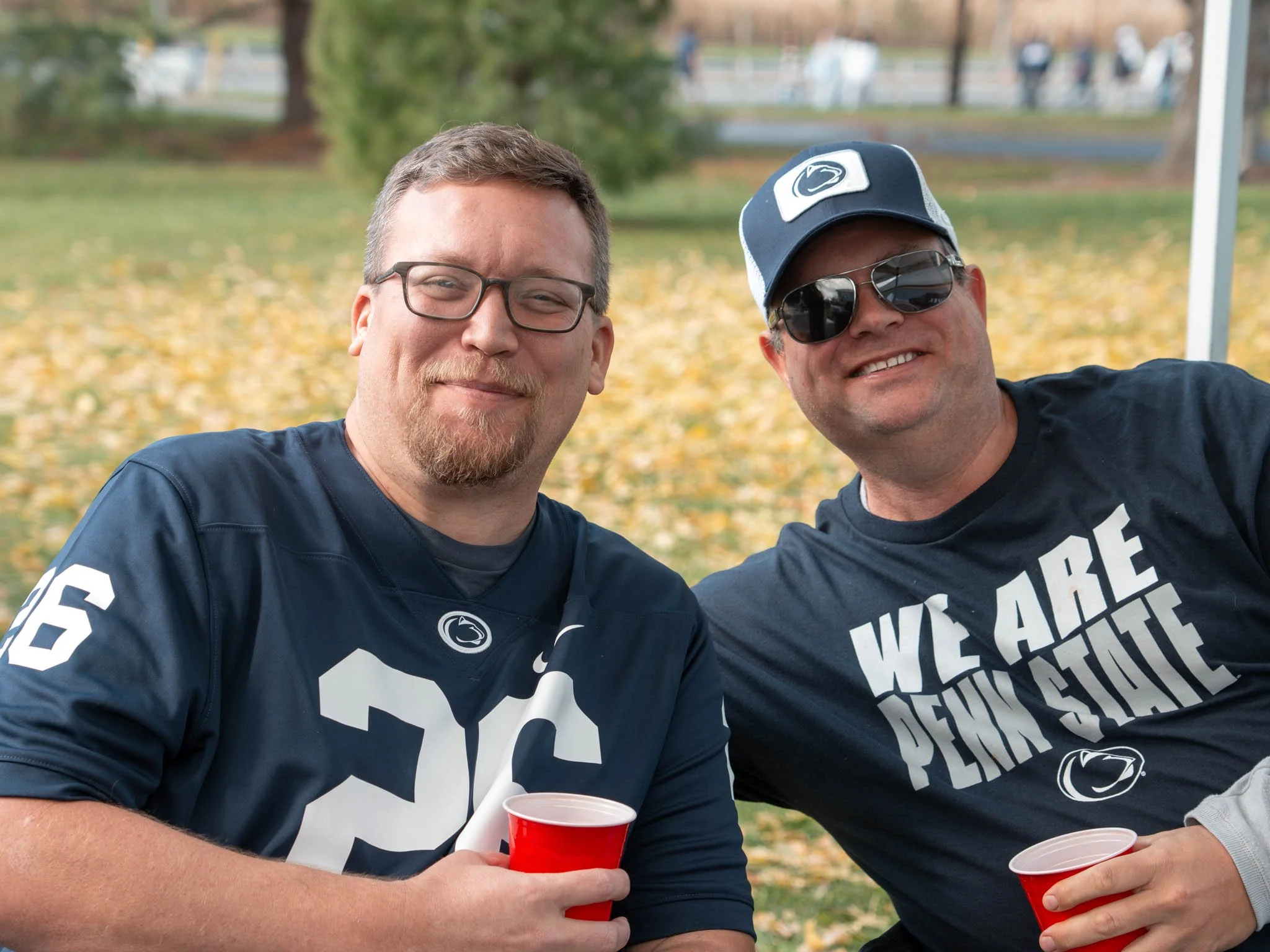 Two men smiling at an outdoor gathering, holding red plastic cups. One wears a Penn State football jersey and glasses, the other wears sunglasses, a cap, and a Penn State shirt that says 'We Are Penn State'. Fall leaves cover the ground in the backgr
