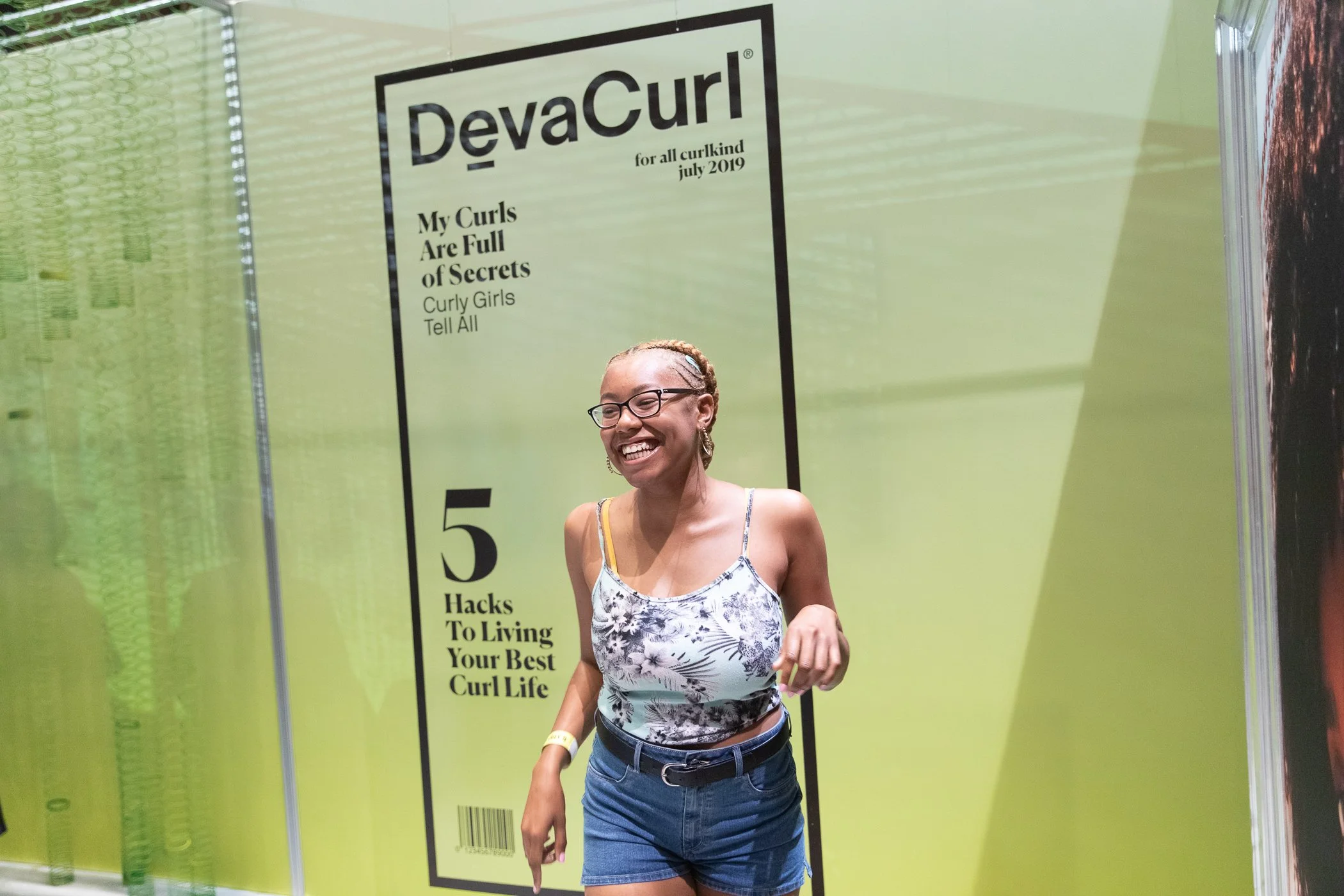 A young woman with glasses and braided hair, wearing a floral tank top and denim shorts, smiling and walking in front of a large yellow poster that reads 'DevaCurl'.