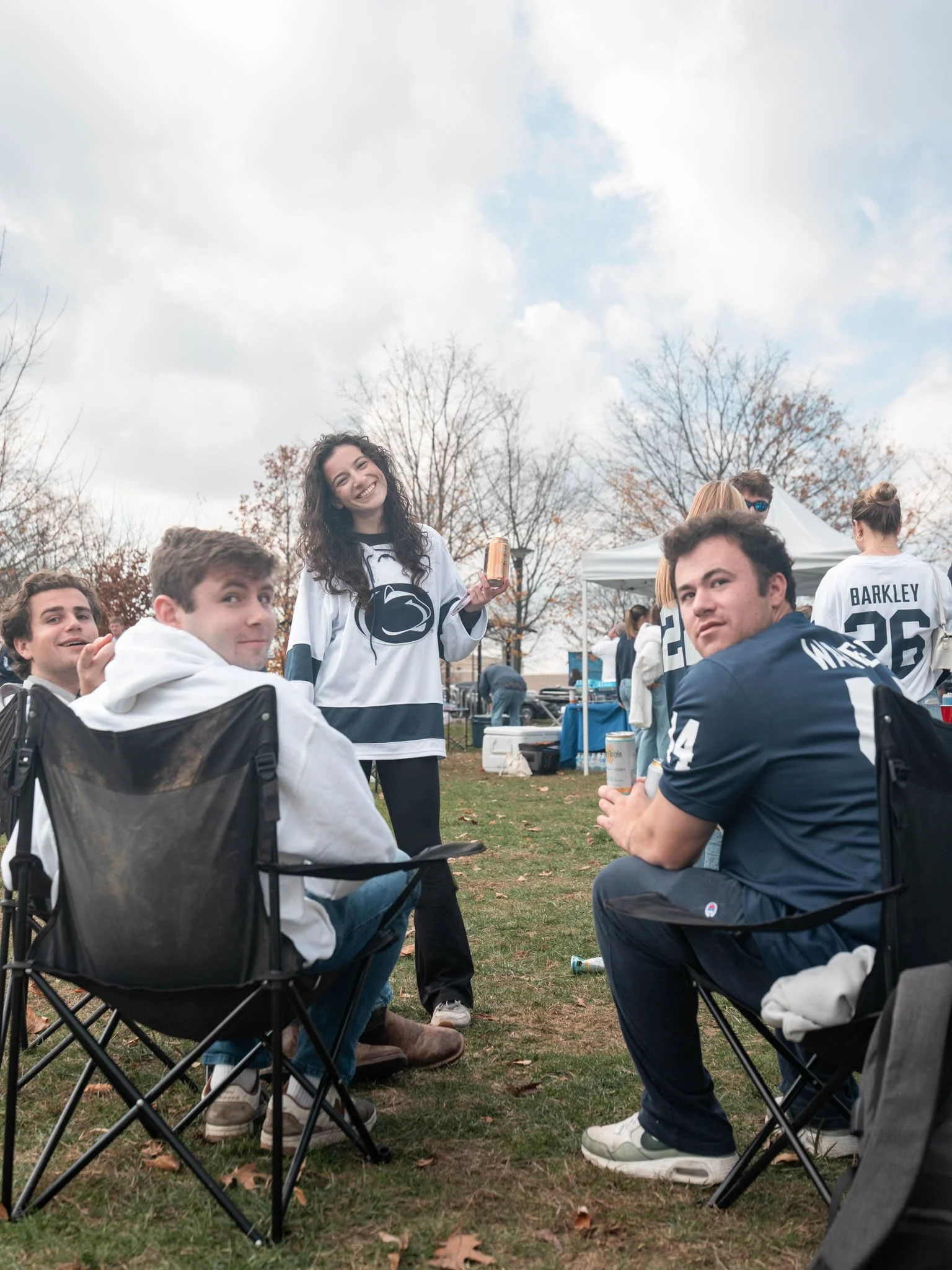 Group of friends at an outdoor tailgate, some sitting on camping chairs, wearing Penn State football jerseys, with a woman standing holding a beverage, smiling at the camera, under a cloudy sky with trees in the background.